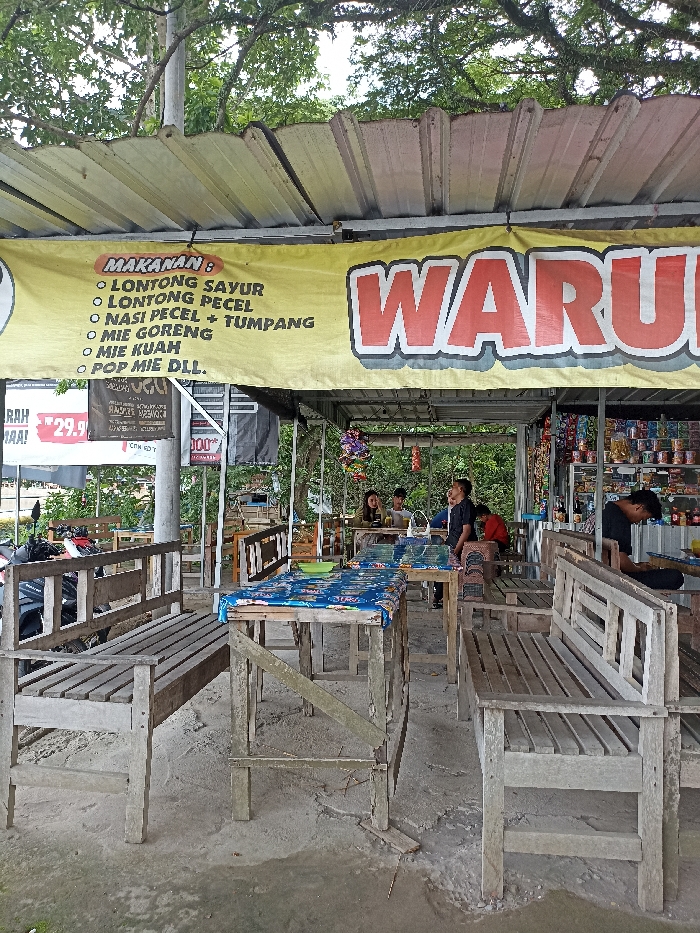 This image captures a casual outdoor eatery, or "warung," situated in Kepung, Indonesia. The setting features a simple structure with a corrugated metal roof, supported by posts, providing shade. Lush green foliage and trees are visible overhead and in the background, suggesting a natural environment. The ground beneath the seating area is a mix of dirt and concrete.
The warung is furnished with rustic, unpainted wooden tables and benches, some tables adorned with blue checkered plastic tablecloths. Several people, appearing to be customers, are seated at these tables in the mid-ground, engaging in conversation or using their phones, contributing to a relaxed atmosphere. Towards the right, a man in a black shirt stands behind a counter, possibly serving or preparing food, while shelves behind him display various packaged snacks and drinks, indicating the establishment also functions as a small convenience store.
A large yellow banner prominently displays the menu items, headed "MAKANAN:" (Food:). The list includes traditional Indonesian dishes such as "LONTONG SAYUR," "LONTONG PECEL," "NASI PECEL + TUMPANG," "MIE GORENG," "MIE KUAH," and "POP MIE DLL." (Pop Mie and others). Large red lettering on the right of the banner spells out "WARU," likely part of "WARUNG." Additional visible text includes "RAH 29.9," "CINDY," "000," and "RASPES MESMOCA" on smaller signs. The scene is illuminated by natural light, indicating it is daytime, and the overall impression is one of a laid-back, local dining spot.