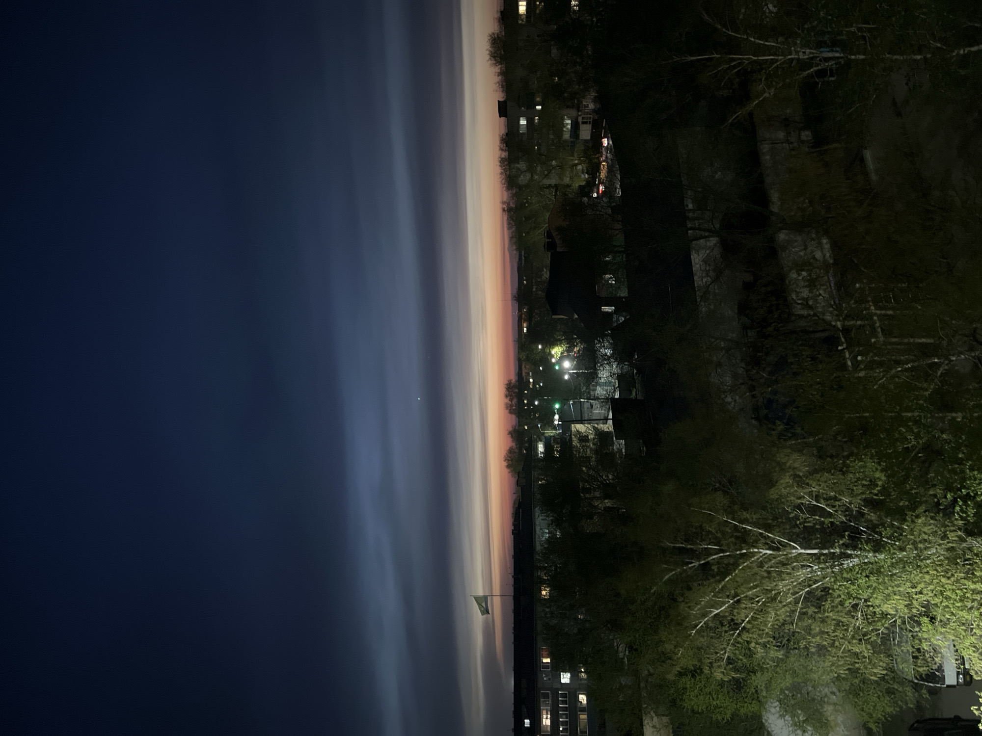 The image depicts a night scene in an urban area, possibly the city of Unknown, Unknown. The sky transitions from a deep blue on the left to a lighter gradient with hints of orange and pink towards the center. This suggests twilight or dawn. Clouds are stretched and blurred horizontally, indicating either long exposure or movement. A flagpole with a flag, possibly Ukrainian, is visible in the middle ground.
Buildings with lighted windows are scattered throughout the scene, with trees framing the foreground and right side. Streetlights illuminate the area.