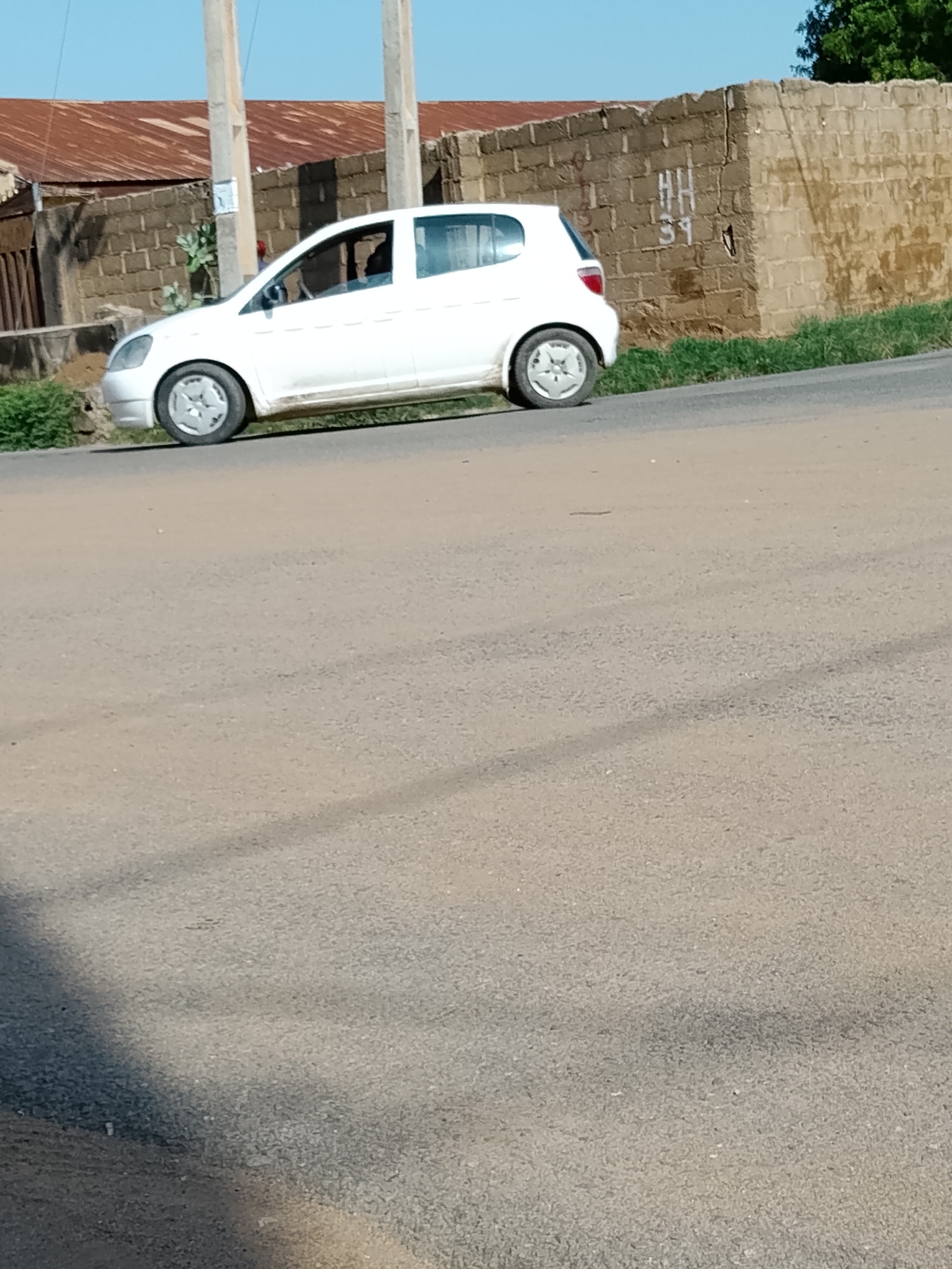 A white compact car is parked on the side of a paved road in Wauro Leggal, Nigeria. The car is facing towards the right of the frame, with its front left wheel slightly turned. Behind the car, a wall constructed of light-colored bricks is visible. The wall has some markings on it, appearing to be letters and numbers spray-painted in white. A corrugated metal roof of a building is visible above the brick wall. Green grass and some small plants grow along the side of the road next to the wall. The sky is clear and blue. The road surface is paved and appears dusty.