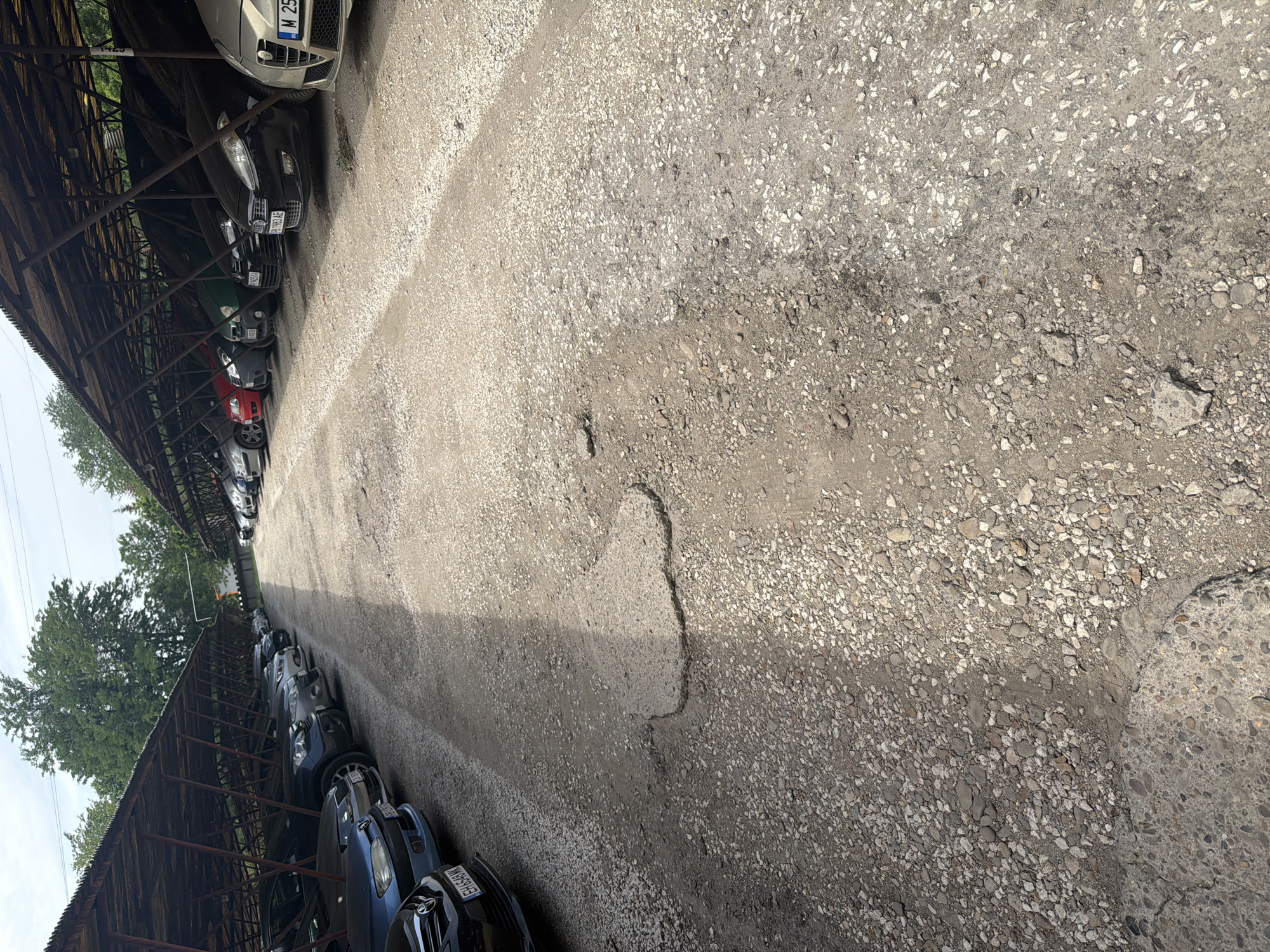The image depicts a long, narrow outdoor parking area in Dolna Mitropolia, Bulgaria. Rows of parked cars are lined under a dilapidated, aged, wooden structure on the left. The ground is an uneven, worn, aggregate-covered surface. The foreground is a close-up view of this cracked pavement. Trees and partial sky are visible on the left further along the row of cars. The photograph has a slight tilted perspective. No people are visible.