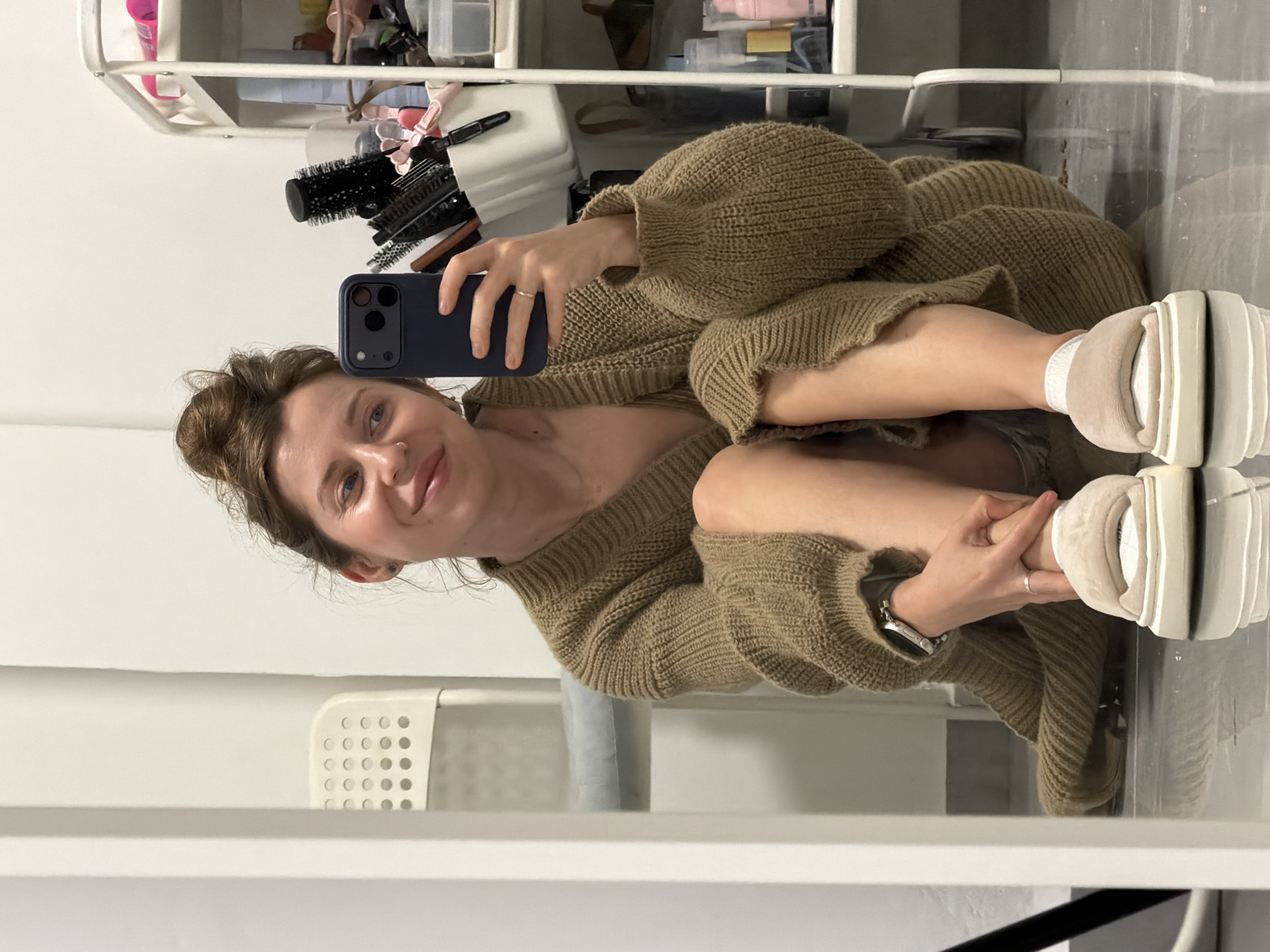 A woman sits on the floor, taking a selfie in front of a mirror. She is wearing a large, tan-colored knit sweater and white platform sneakers with tan trim. She holds a dark blue smartphone in her left hand. The woman has fair skin, light brown hair pulled back in a bun, and a small nose ring. Her left wrist has a silver watch. Behind her is a white wall with a white shelving unit holding various objects, including hairbrushes and containers. A white plastic chair is also visible in the background. The reflective surface suggests a bathroom or dressing area. There is no verifiable information that can confirm this image was taken in the city of Bat Yam, Israel.