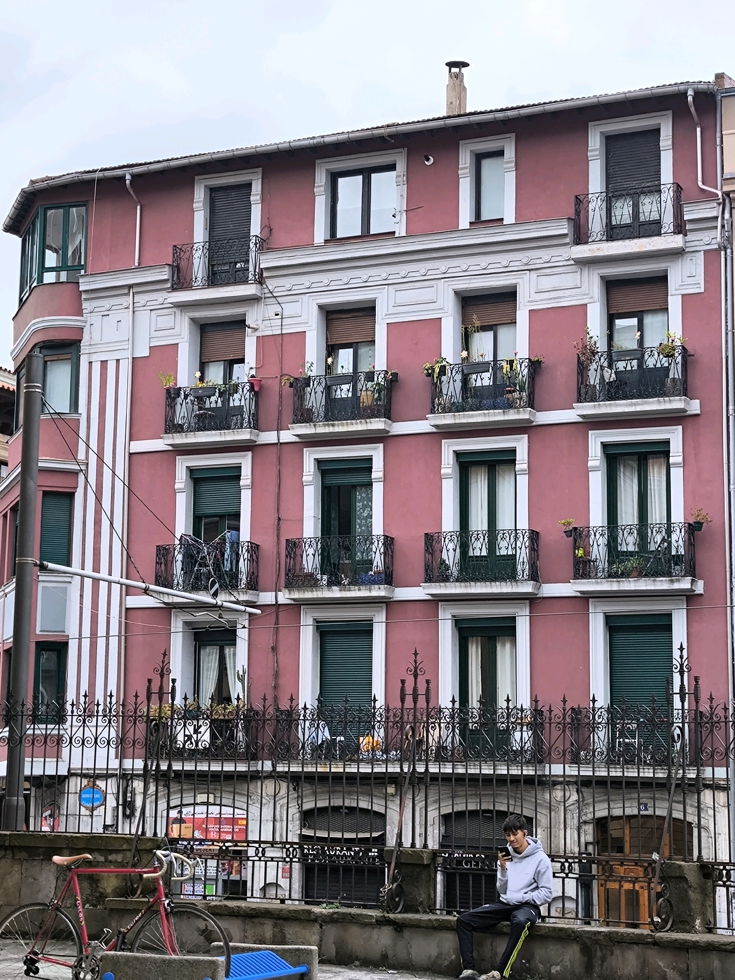 The image depicts a street scene in Bilbao, Spain, focused on a multi-story pink building with white architectural details and numerous balconies. The building features dark green shutters on many of its windows. A black wrought-iron fence runs along the foreground, separating the building from the street. In front of the fence, to the left, a red bicycle is parked. To the right, a young male is seated on the stone wall in front of the fence, looking at his phone. The street itself is not clearly visible, but posters are adhered to the wall behind the fence. The sky is overcast.