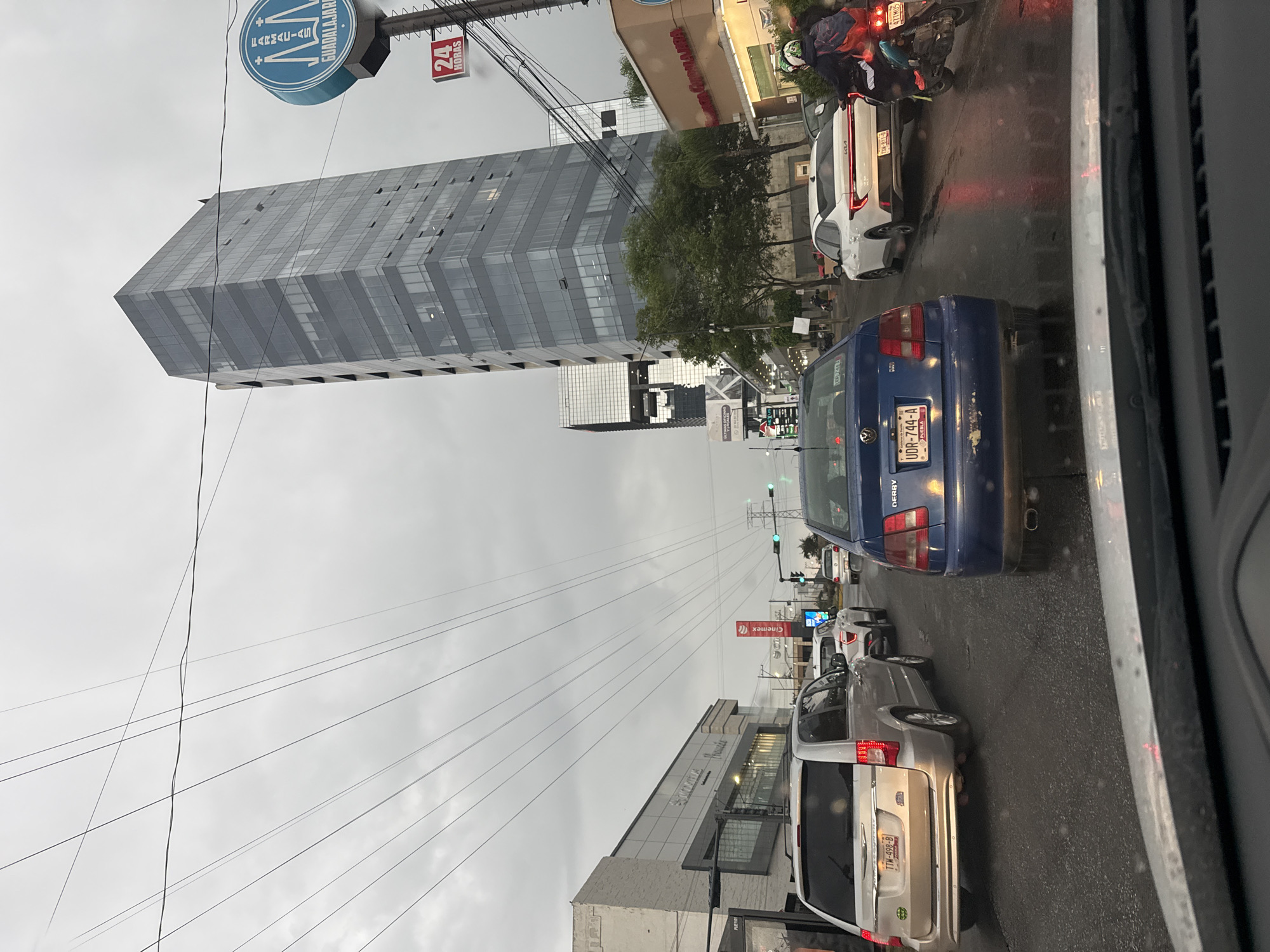 The image, likely taken from inside a vehicle, shows a street scene in Puebla, Mexico on an overcast day. The street is lined with buildings, including a Cinemex movie theater and a Farmacias Guadalajara pharmacy. A tall, modern skyscraper dominates the background. Several vehicles, including cars, vans, and motorcycles, are visible in traffic. A blue Volkswagen Derby has a license plate visible "UDR-744-A" from Puebla. A silver minivan also has a visible license plate "TTW-498-B". Power lines stretch across the sky. The scene suggests urban congestion.