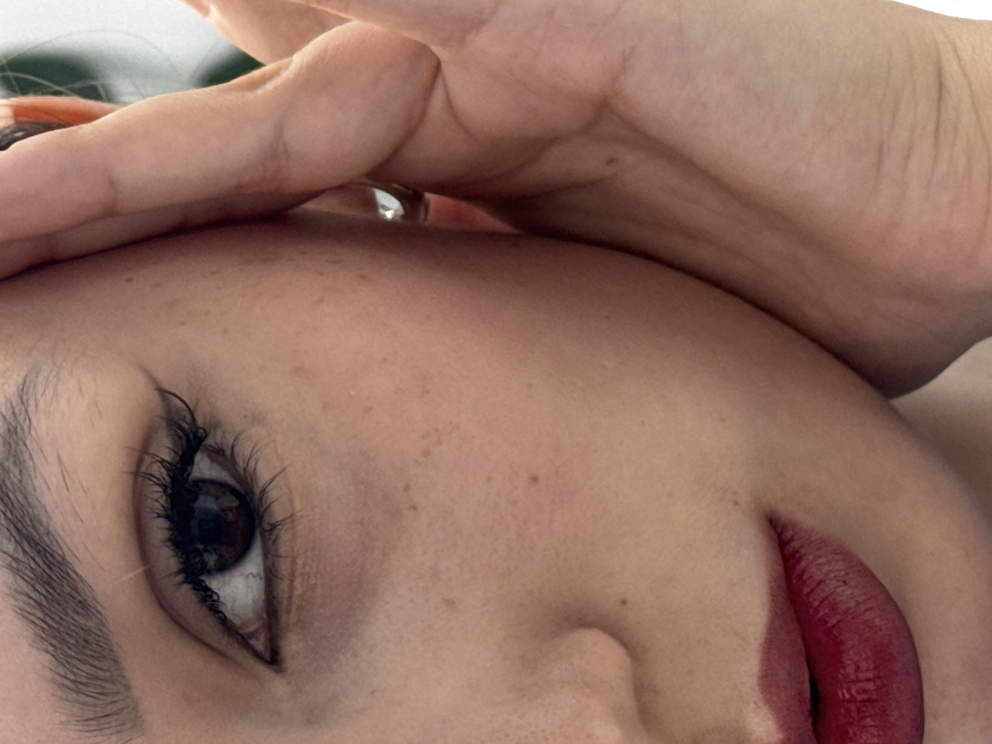 A highly cropped, close-up portrait features a person's face, tilted to the viewer's left. The central focus is on a dark brown eye with prominent, long black eyelashes and a thick, dark eyebrow. Below the eye, the cheek and a portion of the nose are visible, revealing light-toned skin dotted with numerous freckles and small moles. The lower right of the frame showcases full lips, meticulously painted with a deep, matte red lipstick, displaying fine texture.
A hand is positioned across the upper part of the face, resting on the forehead or temple area, with its fingers partially obscuring the head. A silver-toned ring, set with a clear or white stone, is visible on one finger peeking from beneath the hand. The lighting is soft and even, accentuating skin textures and makeup details without harsh shadows. The background is indistinct and light-colored, suggesting a controlled studio setting. The composition emphasizes specific facial features and the hand in a stylized, artistic manner. No text is visible.