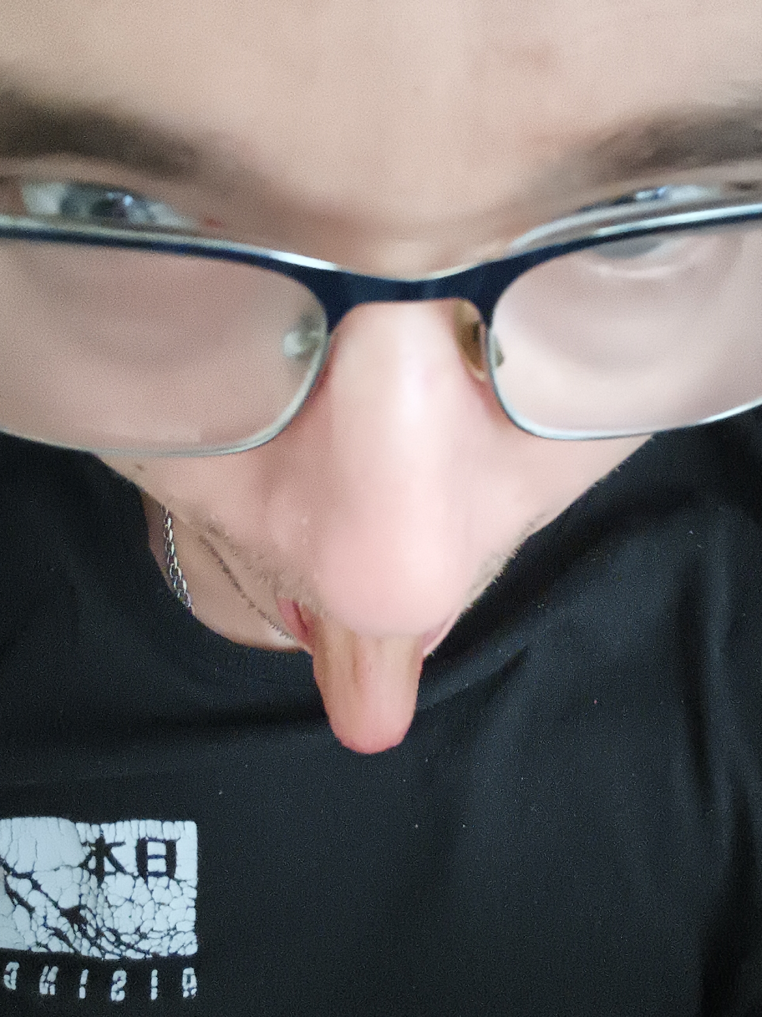 A close-up, low-angle view of a person wearing glasses and a black t-shirt. The focus is on the nose and the area between the eyes. A silver chain is visible around the neck. The t-shirt has a white graphic design of the Japanese word "本" and "日" over a cracked texture, with mirrored numbers below it. The location cannot be determined from the image.