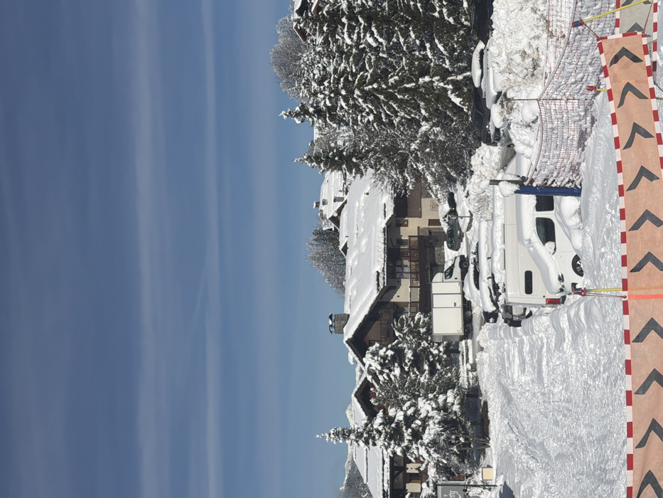 The image depicts a snow-covered village scene in Les Allues, France, under a clear blue sky with faint horizontal cloud formations. The foreground features a wide snow-covered path or access road with visible tracks, bordered on the right by a parking area where multiple vehicles, including a white commercial van, are heavily laden with snow. A patterned safety barrier with orange and white chevron arrows runs along the bottom edge of the frame, accompanied by a segment of red mesh fencing on the right. In the midground, several multi-story chalets or buildings, constructed with stone bases and wooden upper levels, are visible, their dark roofs heavily blanketed in snow. Balconies are present on some structures. Dense, snow-laden evergreen trees are interspersed among the buildings and vehicles. A white box truck is parked centrally among the buildings. No individuals are present. The environment is a mountainous, winter landscape, indicated by distant snow-capped peaks faintly visible on the left horizon.