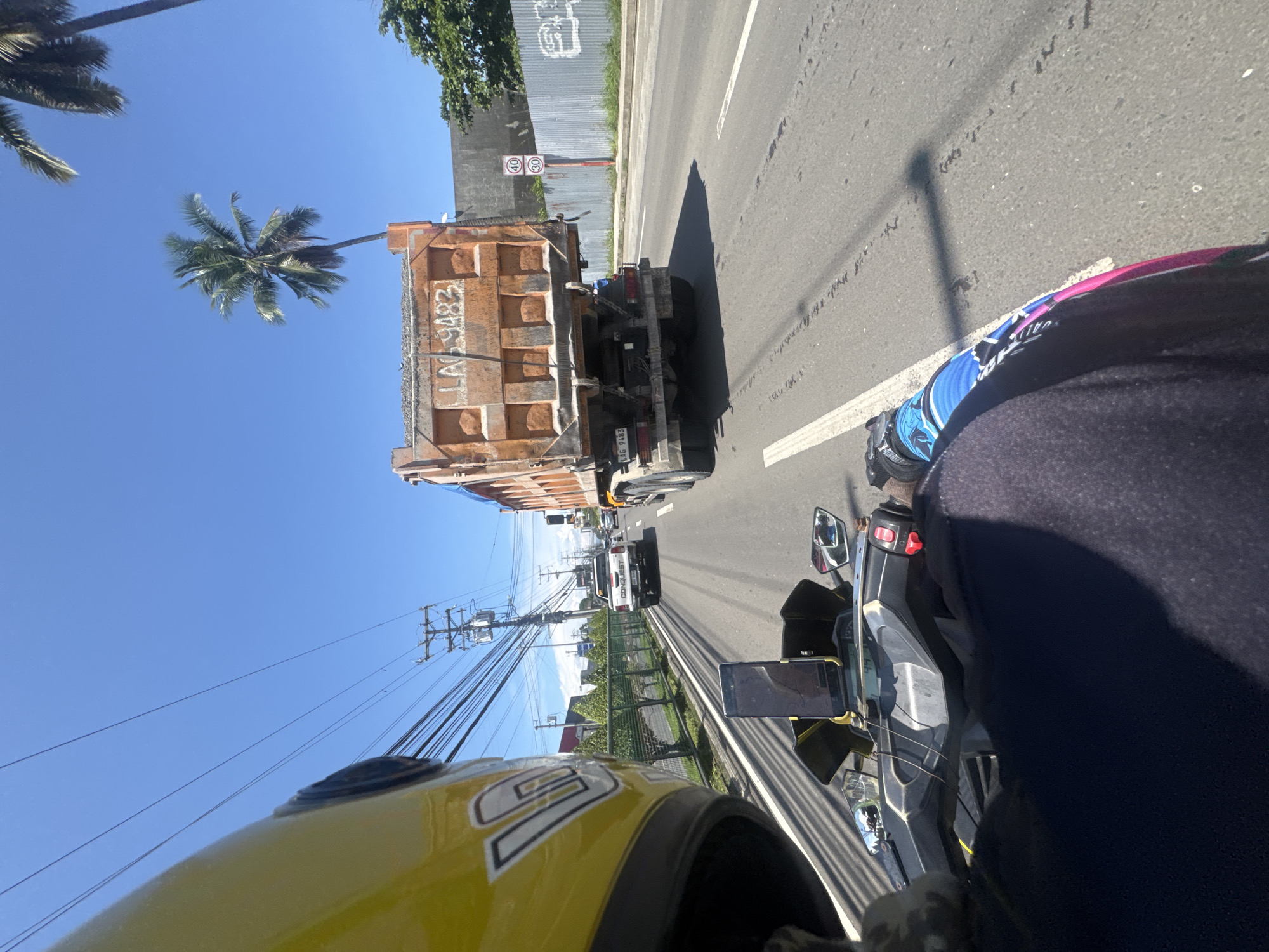 The image shows a view from a moving motorcycle on a road in Davao City, Philippines. A large orange dump truck is directly ahead, followed by other vehicles. A yellow motorcycle helmet is visible in the foreground, with the rider wearing blue and black riding gear. Palm trees and utility poles line the side of the road. A sign with speed limits (30 and 40) is visible near the dump truck. The dump truck's plate number is LAG9483.