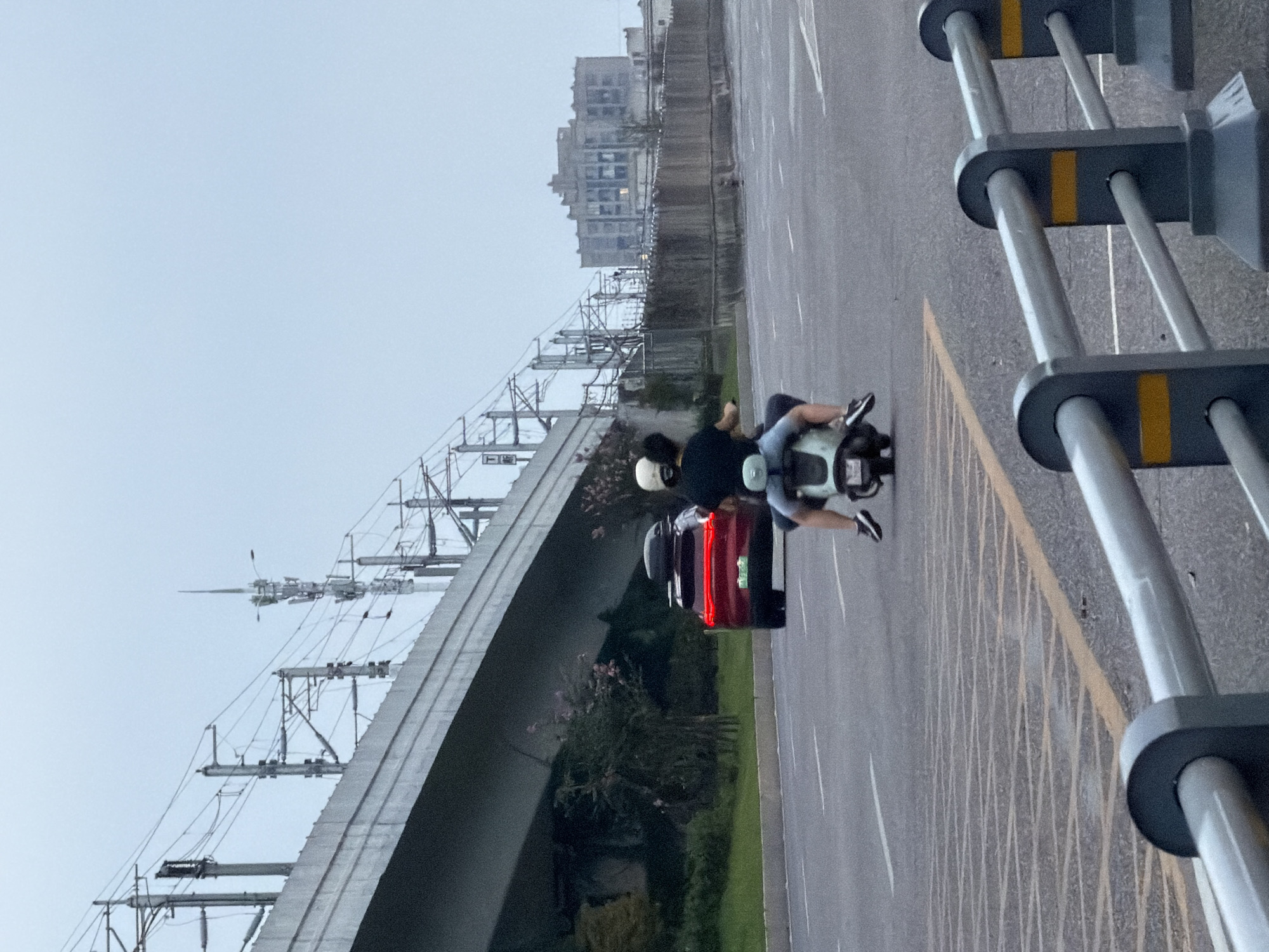 An overcast, late afternoon scene is depicted in Jiangjin, China, featuring a two-person ride on a scooter. The scooter, a light green moped, travels away from the viewer down a paved road. A person in the rear, wearing a white helmet and a dark t-shirt, is visible from the waist up. They are sitting behind the driver, who wears a dark helmet and a dark t-shirt with light blue denim shorts.
In the background, a red SUV with a roof rack is in the left lane, also moving away from the viewer. The vehicle has illuminated brake lights and a visible license plate. Further in the distance, a complex network of overhead power lines and utility poles spans across the sky, suggesting proximity to a transportation or industrial area. A long, elevated concrete structure runs horizontally across the upper left quadrant of the image, likely an overpass or bridge.
To the right of the road, a tall, gray, multi-story building with visible windows stands behind a wooden-slatted fence. The road itself is marked with white lane dividers and a yellow painted diagonal pattern in the foreground, indicating a potential traffic calming zone or pedestrian crossing area. A metal guardrail with gray supports and horizontal silver bars, accented with yellow bands, is positioned in the foreground, partially obscuring the lower portion of the scene. The overall lighting suggests a diffused, possibly rainy or cloudy, atmosphere.