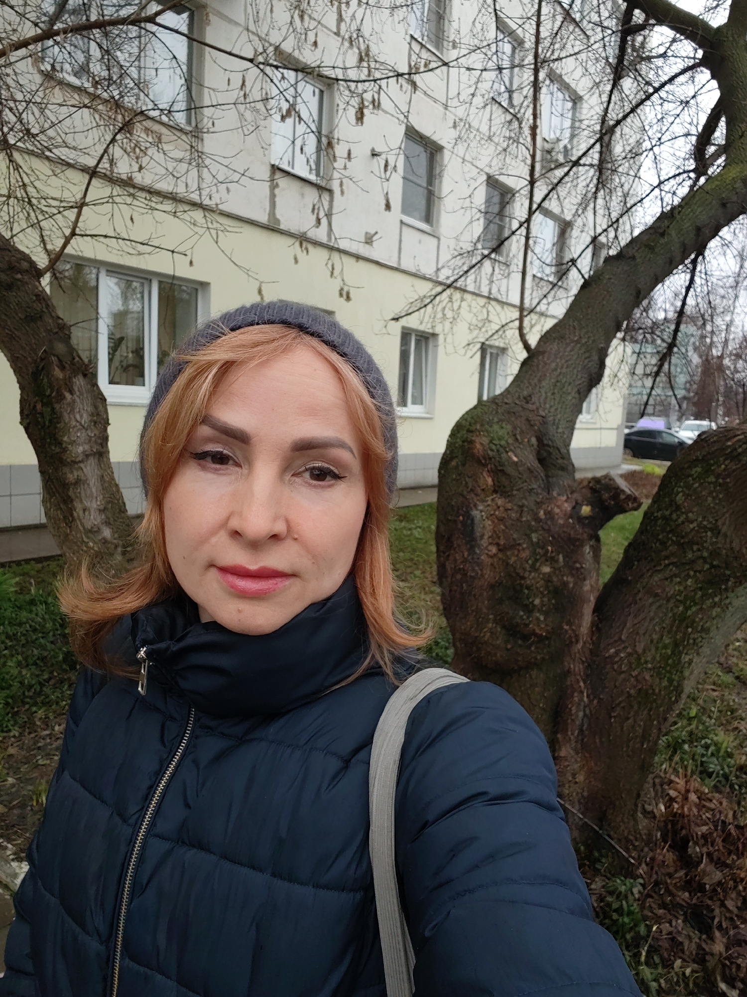 A woman in a dark blue puffer jacket and gray hat stands outside in front of a beige building with white-framed windows. The building appears to be an apartment complex. To the right of the woman is a bare tree with a thick, gnarled trunk. A silver strap crosses her shoulder. In the background, cars are visible on the street. The location is Chaikovsky, Russia.