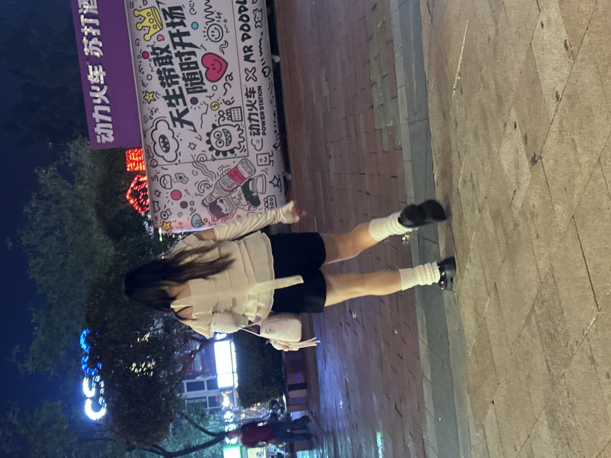 A woman walks away from the camera at night on a brick sidewalk. She has long dark hair and is wearing a white off-the-shoulder top with puffy sleeves, black shorts, white thigh-high socks, and platform shoes. She carries a white handbag. Behind her, a large purple sign with Chinese characters and cartoon drawings is visible. A blue neon sign with the letters "CSG" is also visible in the background. The setting appears to be a commercial or entertainment area. The ground is wet, suggesting it has recently rained. The woman is the primary subject, and the environment is an outdoor urban setting at night.