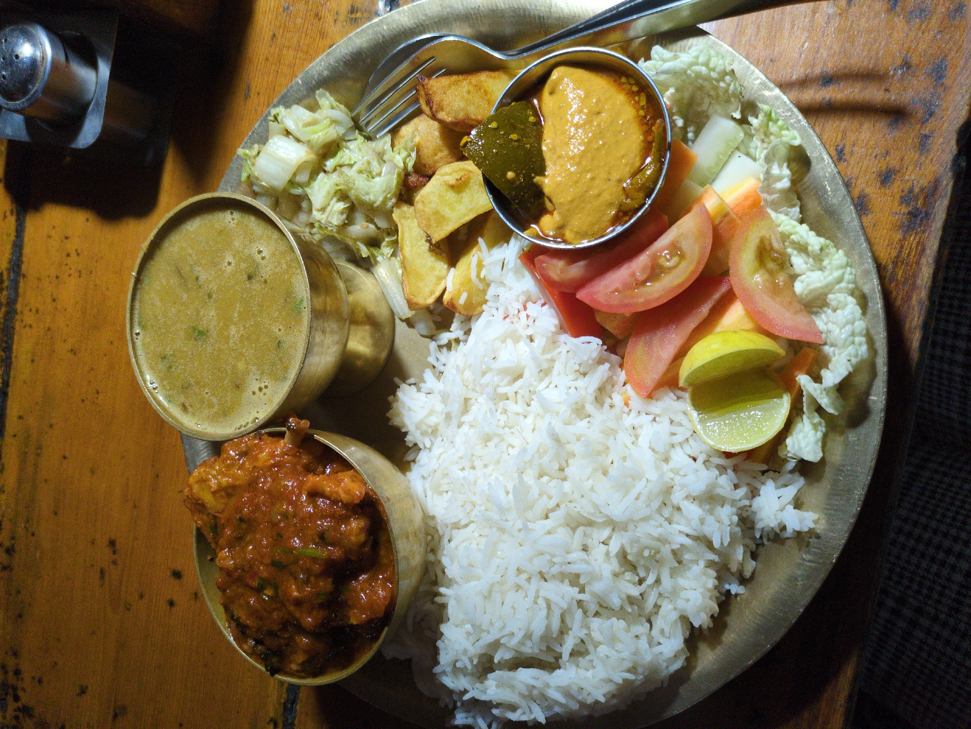 An overhead shot captures a traditional Indian meal served on a brass thali, placed on a wooden table. The thali contains a generous portion of white rice, a bowl of reddish-brown curry with visible meat pieces, a smaller bowl of pale green soup, and a side of chopped vegetables, including tomato slices, cucumber sticks, and shredded cabbage. A small metal bowl holds a yellowish dip, possibly a chutney, with a piece of green chili submerged in it. Fried potato wedges are scattered around the thali. A fork rests on the right side of the plate. In the background, a metallic salt and pepper shaker set is visible on the wooden table, with a dark shadow cast behind it. The overall scene suggests a meal served in a restaurant setting in Bhagsu, India.