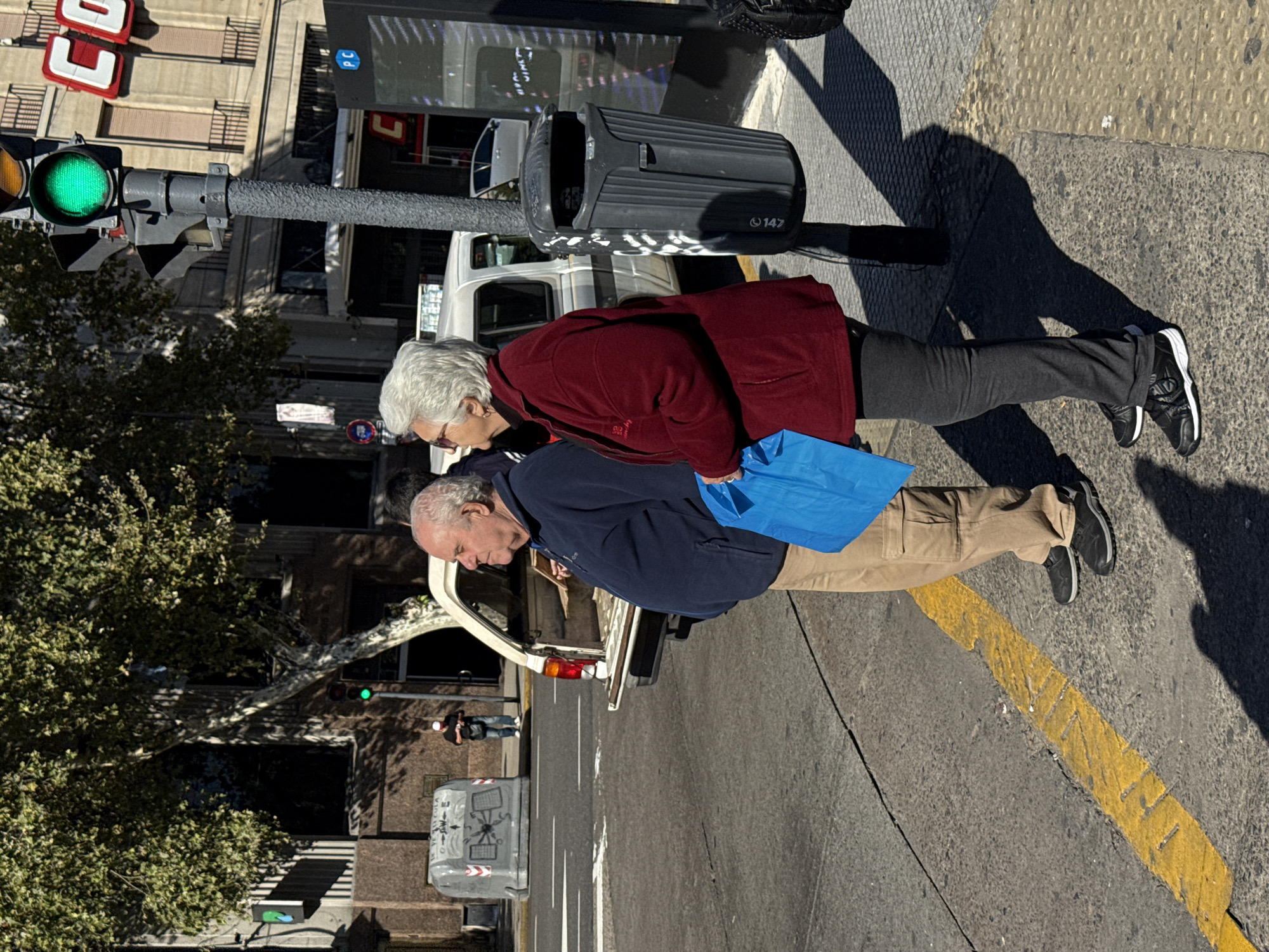 An elderly couple is standing on a sidewalk in Buenos Aires, Argentina. A man is wearing a navy jacket, khaki pants, and black shoes. A woman is wearing a maroon jacket, gray pants, and black shoes. They are carrying a blue shopping bag. They are next to a car parked on the street. The traffic light is green. The city is well lit by the sun.