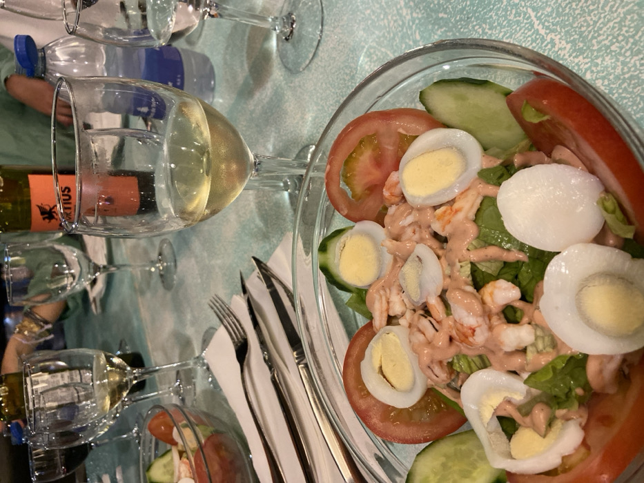 The image shows a table setting in a restaurant located at 28.738, -13.867 in Corralejo, Spain. A glass of white wine is partially filled, sitting next to a glass of water and a bottle of white wine. The table has a light blue tablecloth. A glass bowl filled with salad is in the foreground. The salad contains shrimp, lettuce, tomato, cucumber, and hard-boiled eggs. The salad is dressed in a creamy pink sauce. There are two forks and a knife on the table. A white napkin is partially visible behind the salad bowl. The table is partially visible in the background.
