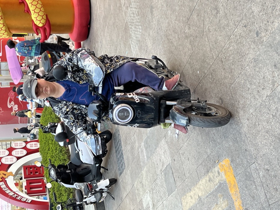 The image portrays an outdoor urban environment in Jiangjin, China, under bright daylight. In the foreground, a person of stout build is seated on a black electric scooter. This individual wears a dark grey baseball cap with a white Nike logo, a long-sleeved jacket patterned with black, white, and grey designs, a blue t-shirt, dark blue sweatpants, and maroon flat shoes. They are positioned facing the camera, with a neutral facial expression, gripping the handlebars of the black scooter. The scooter features a circular LED headlight and the alphanumeric identifier "009" on its front panel and below the handlebars. Transparent handguards are attached to the handlebars.
To the left in the mid-ground, two additional electric scooters are parked: a white one with a black seat and rear storage box, and a smaller white scooter. Behind these vehicles, a line of green shrubbery precedes a large red architectural archway decorated with white Chinese characters. The ground in this area is paved with grey rectangular stones, and a short yellow painted line is visible in the lower-left corner.
In the mid-ground to the right, a large, cylindrical, red and gold decorative structure, resembling a segmented dragon, stands prominently. Several other individuals are visible in the background, out of focus. One person, wearing a colorful patterned jacket, walks past the dragon decor, while another holds a pink umbrella. A small market stall with items displayed is partially visible further back. The overall environment suggests a bustling commercial or public plaza.