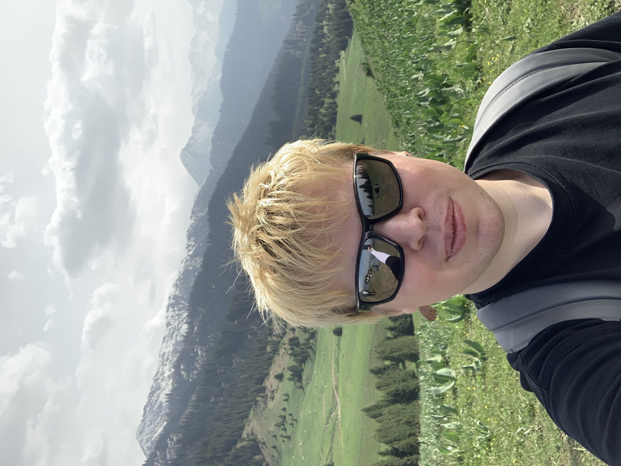 A medium shot depicts a person with short, blond hair wearing dark sunglasses and a black shirt with a gray backpack strap visible. The person is positioned against a backdrop of mountainous terrain. The landscape includes forested slopes and grassy fields. The sky is overcast. The image appears to be taken on a hillside. While the image is potentially located in Almaty, Kazakhstan, there is no specific identifiable feature in the image to confirm the location.