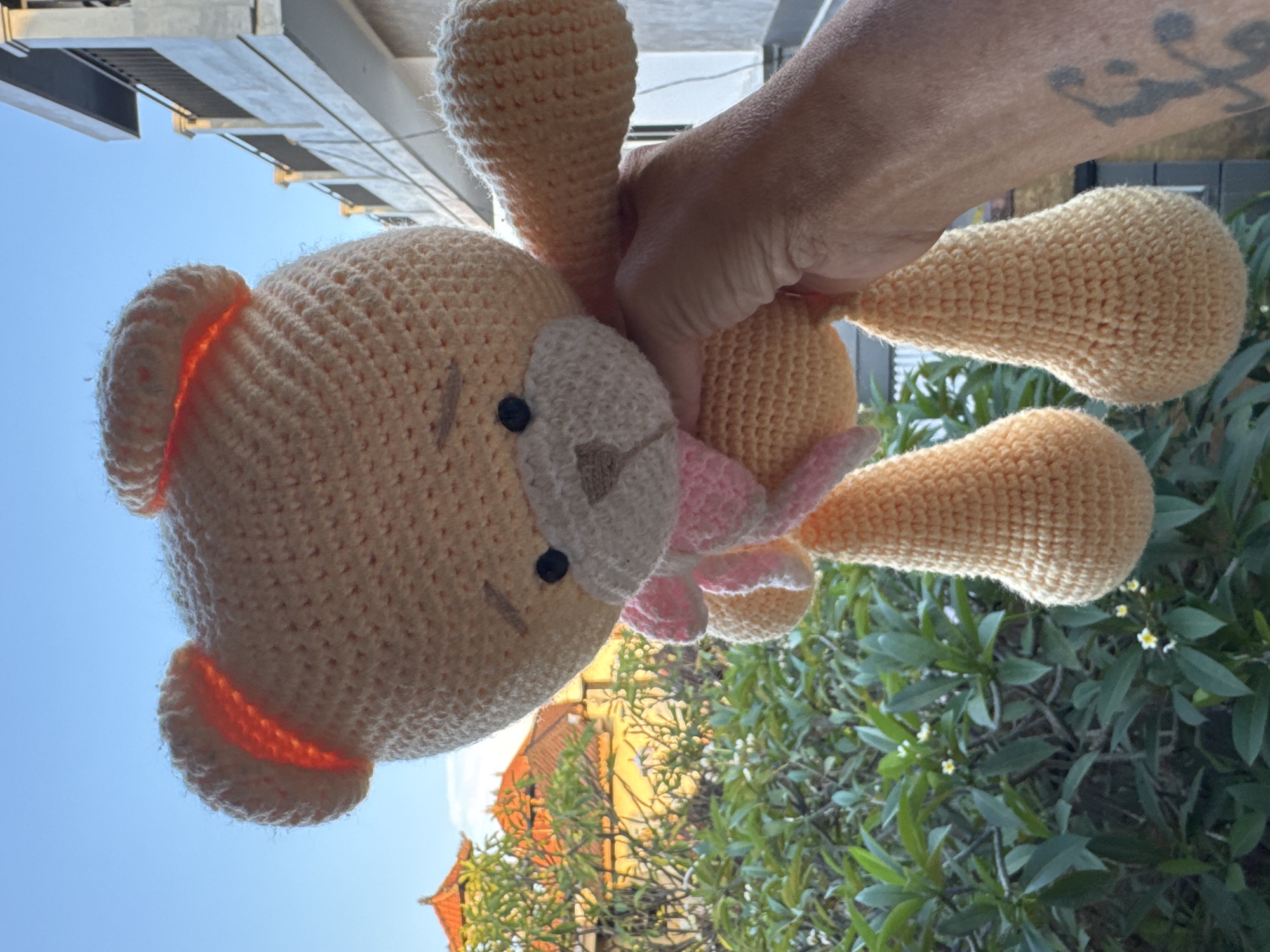 A brightly lit, outdoor image showcases a person's hand holding a crocheted stuffed animal against a vibrant blue sky. The image is rotated 90 degrees counter-clockwise, making the sky appear on the left and ground elements on the right.
The central subject is a light beige/cream-colored crocheted toy, resembling a bear or bunny, featuring large ears and a small pink bow around its neck. It has black eyes and an embroidered light brown nose and mouth. The intricate crochet stitches are clearly visible, indicating its handmade nature. The bright sunlight, positioned behind the toy, creates a warm, orange glow within the translucent yarn of its ears.
A person's hand and forearm are visible, gripping the toy from what would be the top right of the frame if the image were correctly oriented. A prominent dark tattoo, depicting a stylized figure or symbol, is visible on the person's forearm.
The background presents a clear, bright blue sky that dominates the upper and left parts of the frame (when re-oriented). Against this backdrop, modern, multi-story buildings with balconies or windows are visible on the left side of the background. On the right side, dense, lush green foliage with small white flowers covers a significant area, with an orange-tiled roof structure partially visible beneath it. The bright, sunny conditions and tropical vegetation are characteristic of Kuta, Indonesia.
The scene depicts a simple act of holding up a toy outdoors on a clear day. No visible text is present.