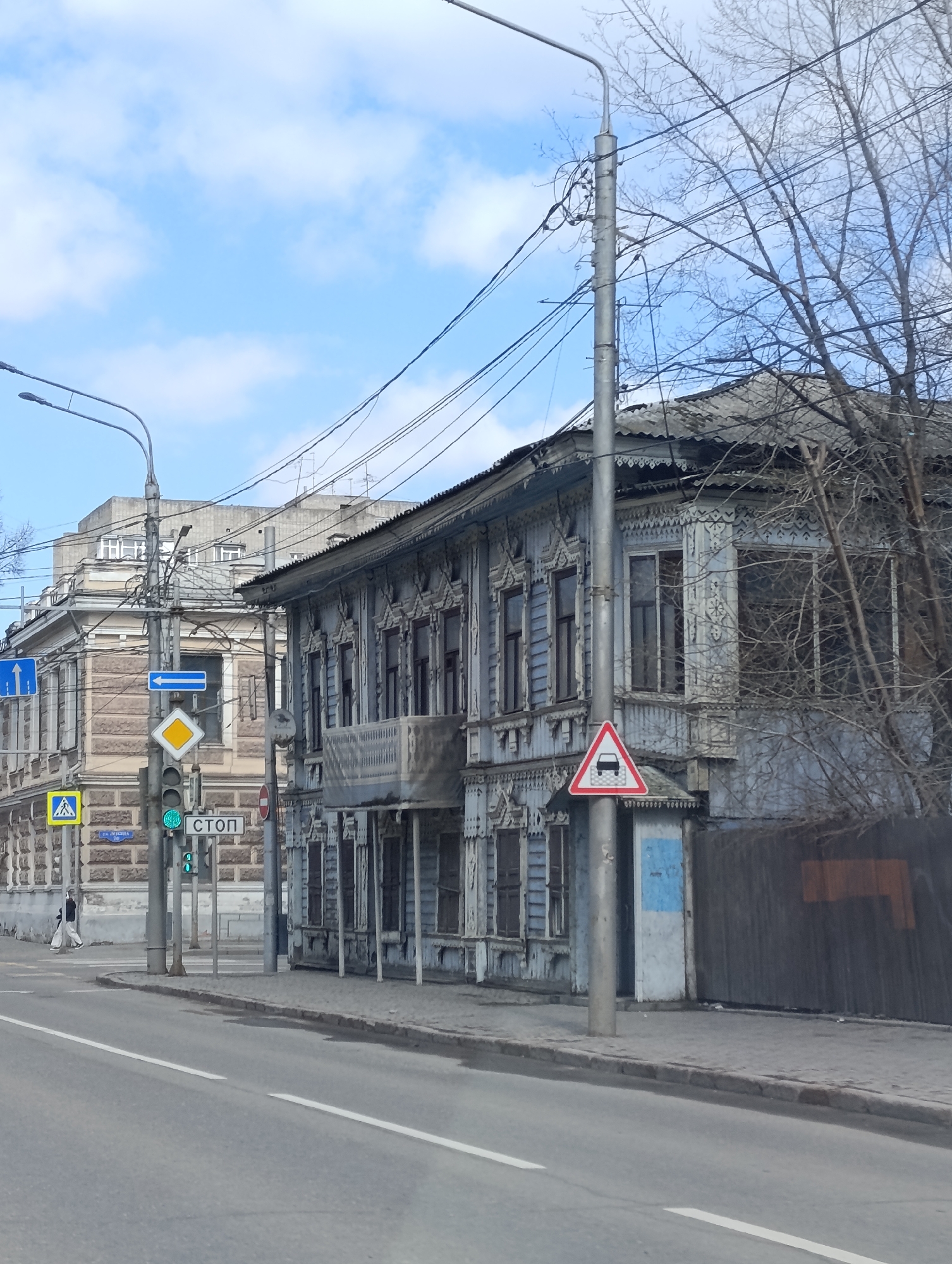 The image shows a street scene in Krasnoyarsk, Russia. The architectural styles suggest the city's urban environment. There is a wooden, two-story building with intricate carved details on the facade next to a traffic pole. The building appears somewhat weathered, indicating age. Adjacent to the wooden building, is a fence, likely marking a property boundary. There is a stop sign in Russian, with the word "СТОП" shown on the street sign. In the background, there is a taller, stone building. A pedestrian is seen crossing at a pedestrian crosswalk. There are traffic lights showing a green light for vehicles. Bare trees stand to the side, suggesting either late autumn or early spring. Power lines are visible overhead, crisscrossing above the street and buildings. The sky is partly cloudy.