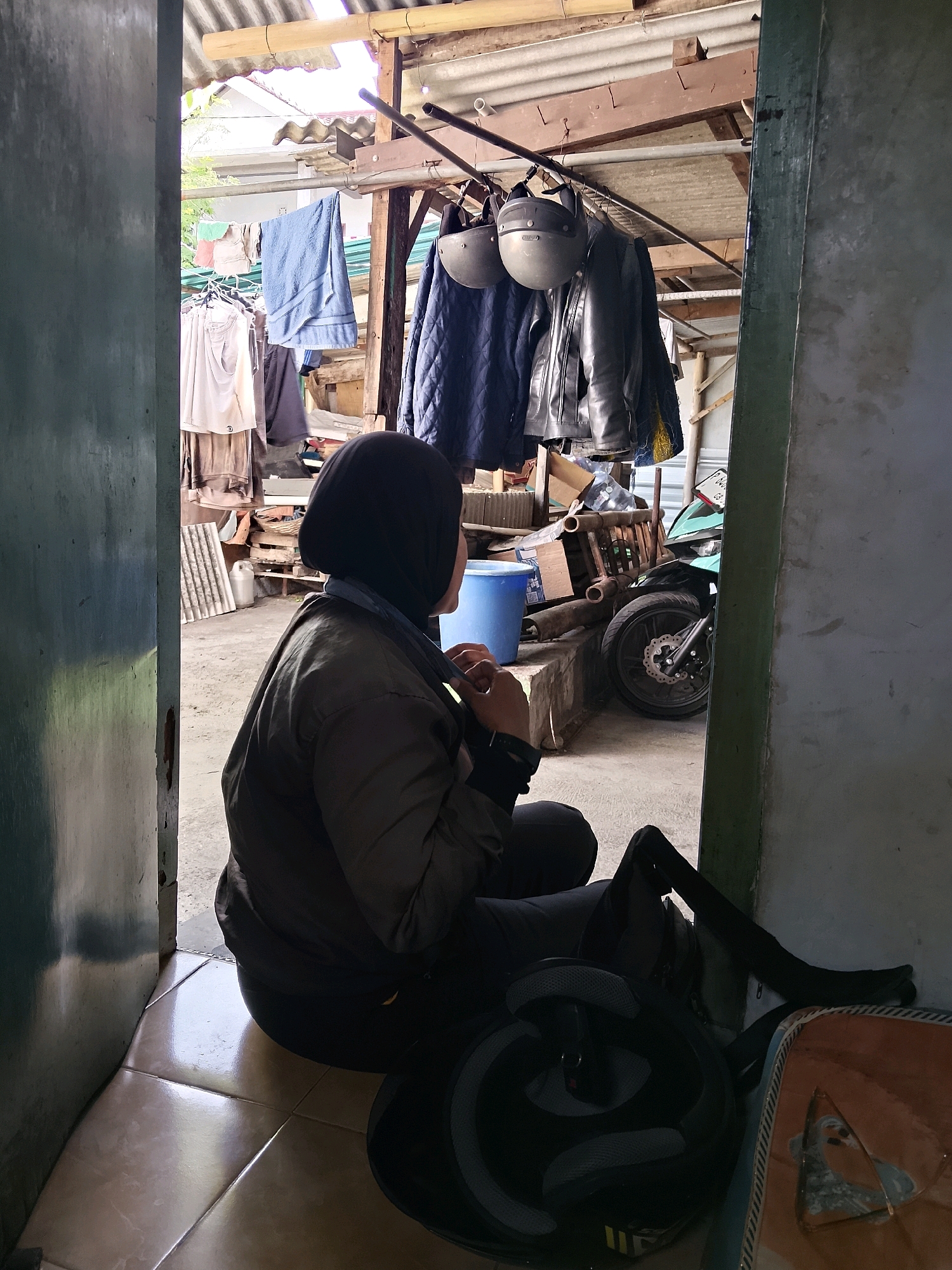 A person wearing a black hijab and a dark olive jacket is sitting on a tiled floor, looking away from the camera and towards a storage area. The person is hunched over, with their hands near their neck. In front of the person, a black motorcycle helmet and a backpack are visible. The background shows a cluttered storage area with various items including clothes hanging on a line, a blue bucket, stacked boxes, and a turquoise motorcycle. The scene appears to be an outdoor or semi-enclosed space, possibly a backyard or garage, with a corrugated metal roof supported by wooden beams. The image has a raw, unfiltered quality, suggesting it was taken candidly. This scene is located in Jember, Indonesia.
