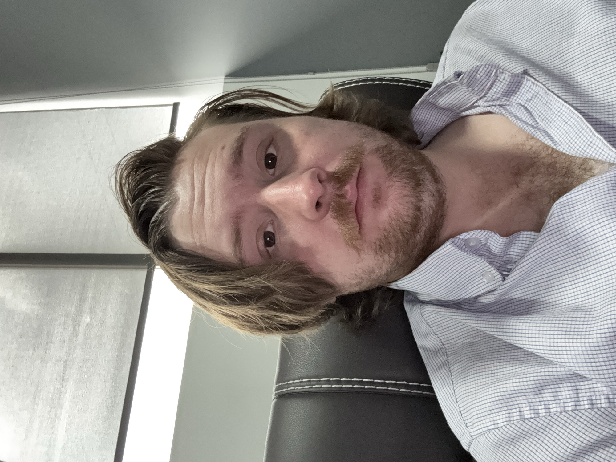 The image shows a fair-skinned man with long, light-brown hair and a beard, wearing a blue and white striped shirt. He is sitting in a black chair with white stitching along the edges. The background includes a grey wall and a window with blinds. The image appears to be taken indoors, possibly in an office or home setting. The location is not discernable from the image.