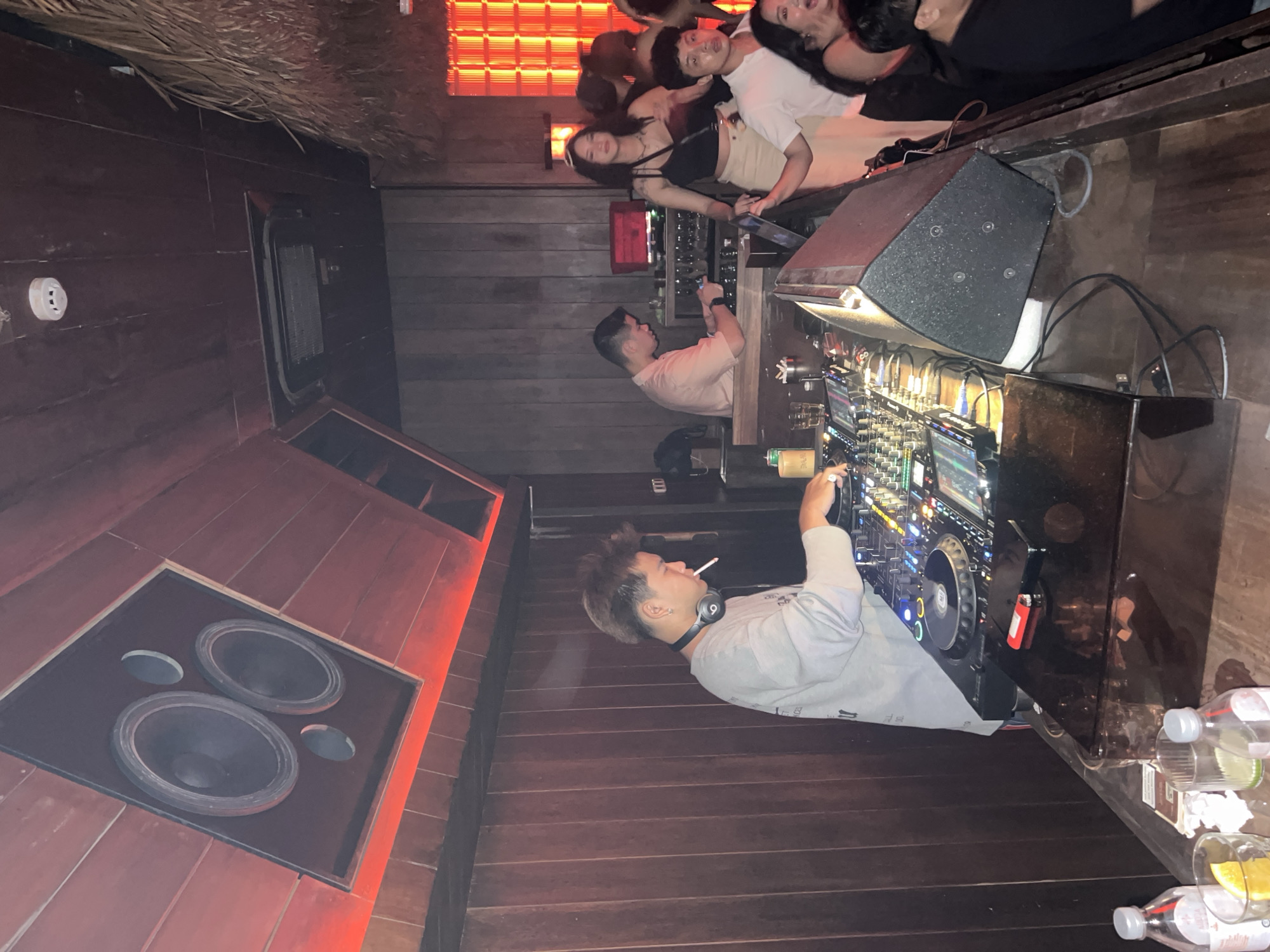 A dimly lit indoor venue, possibly a nightclub, is shown. A DJ booth is the focal point, with a DJ in a grey shirt and headphones visible while smoking. Music equipment is visible on the booth. There are speakers mounted on a wooden wall with red lighting. Several patrons are clustered near the DJ booth. The location is specified as Thủ Đức, Vietnam.