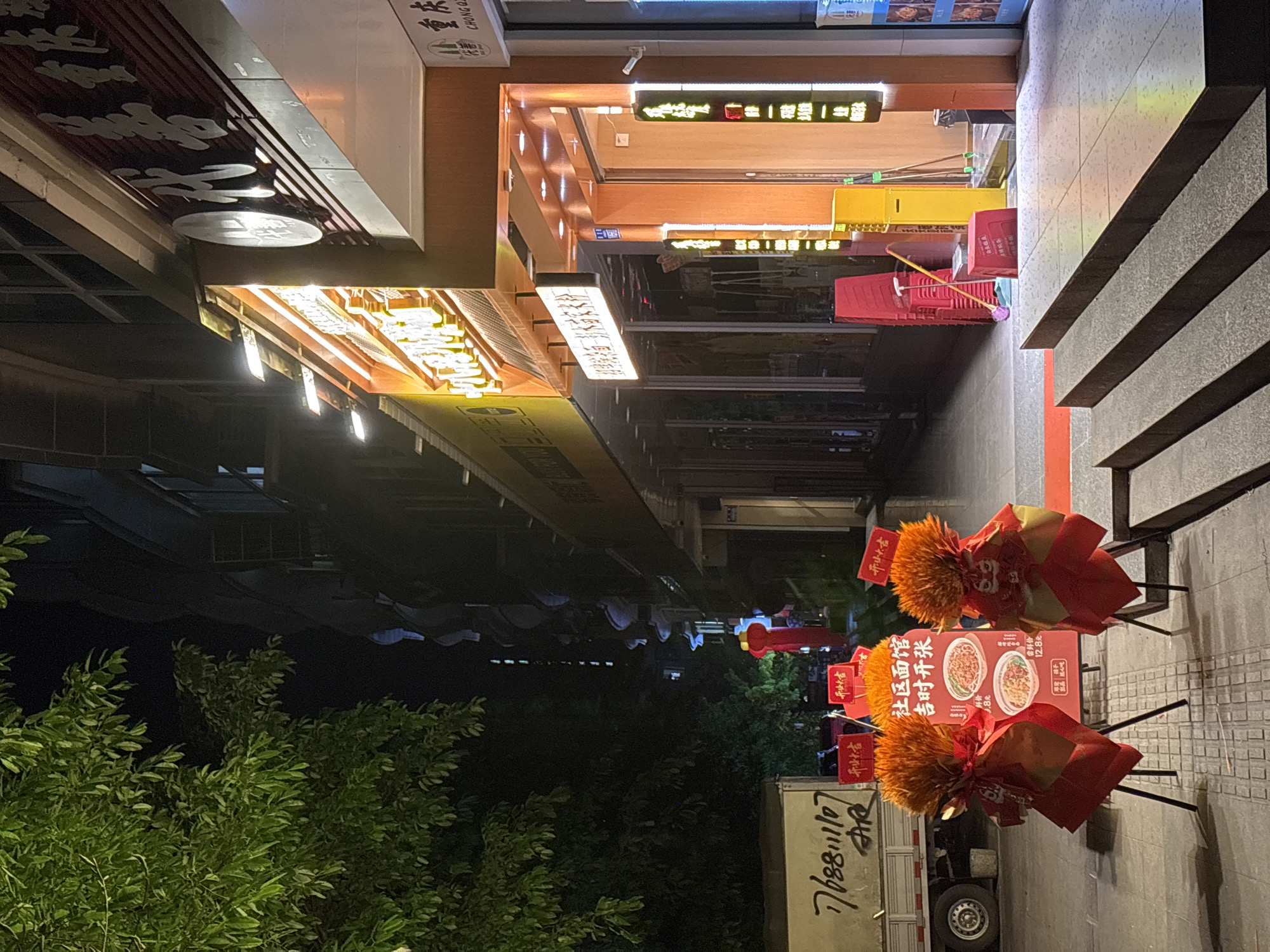This is an outdoor scene taken in Nan'an District, China, at night. The viewpoint is from a street-level perspective looking down a row of storefronts and street art towards the end of the street.
The street is lined with buildings that have brightly lit signage, written in Chinese, under awnings on the left side. The stores have lighted signs that include the names of businesses and products. A few red plastic chairs and other small items are leaning against the buildings on the right side.
The right side of the image depicts a walkway, which has a staircase that runs along the right side. Near the foreground are two decorative promotional display stands. They are covered in red and gold decorations, including a floral-like top and a menu displaying noodle dishes. One stand reads "社区面馆 吉时开张" (Community Noodle Restaurant - Opening at an Auspicious Time), with prices listed for dishes.
To the left and behind the display stands is a partial view of a white truck with painted black numbers, "768811107", in reverse and stylized Chinese script. The truck sits in front of lush, green trees and shrubbery. Farther down the street, the scene is lit with red lanterns strung up along the street.