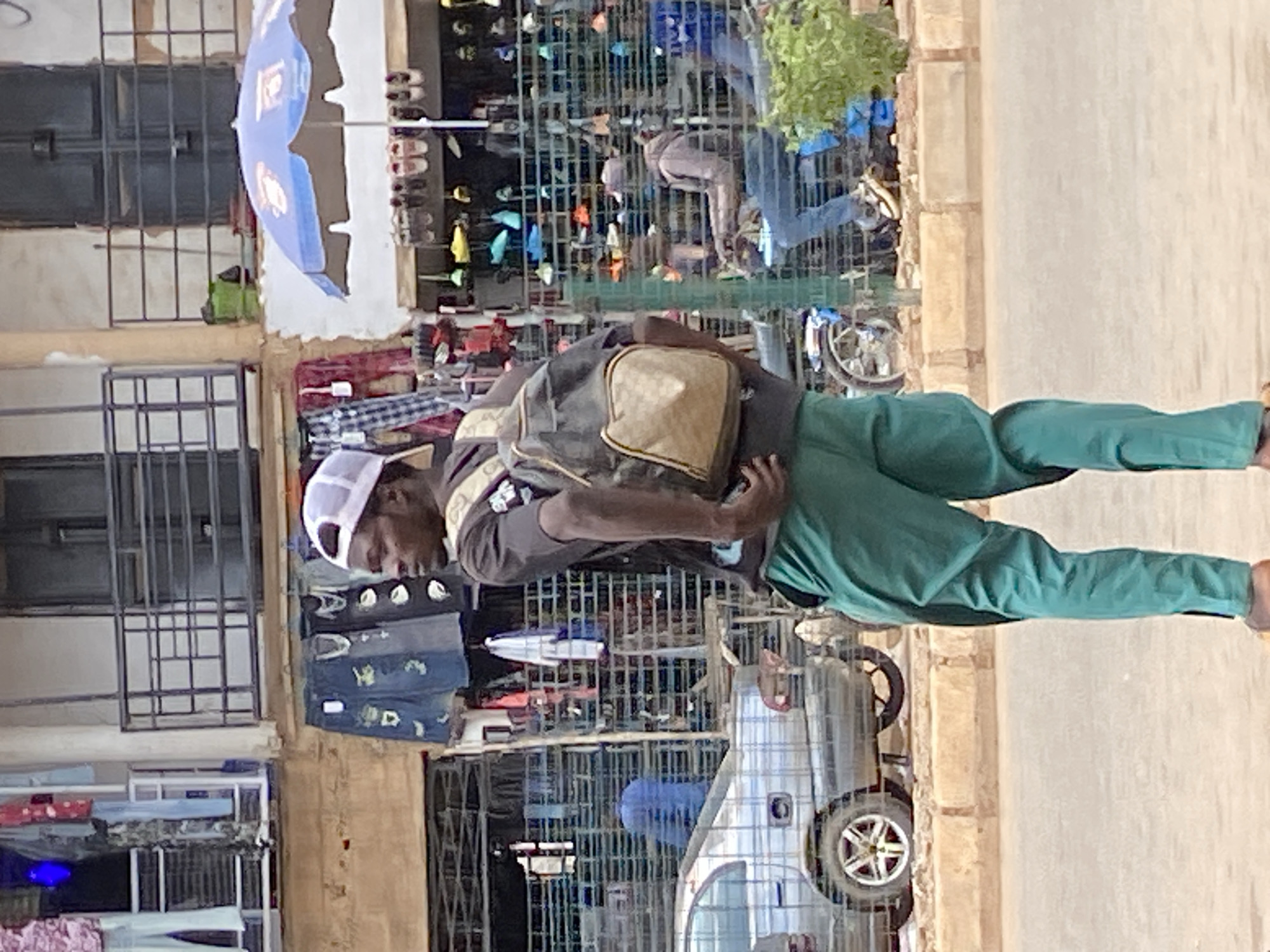 In Bukuru, Nigeria, a person wearing a light-colored cap, a dark t-shirt with a graphic design, and bright green pants walks down a street. They are carrying a dark backpack with a beige accent. The person is walking away from the camera, looking to their left. Behind them, a gray car is parked on the side of the road. In the background, a market is visible with stalls displaying clothing and shoes, and a blue umbrella. Another person is seen riding a motorcycle in the distance, also in a market setting. The scene appears to be a bustling urban environment.
