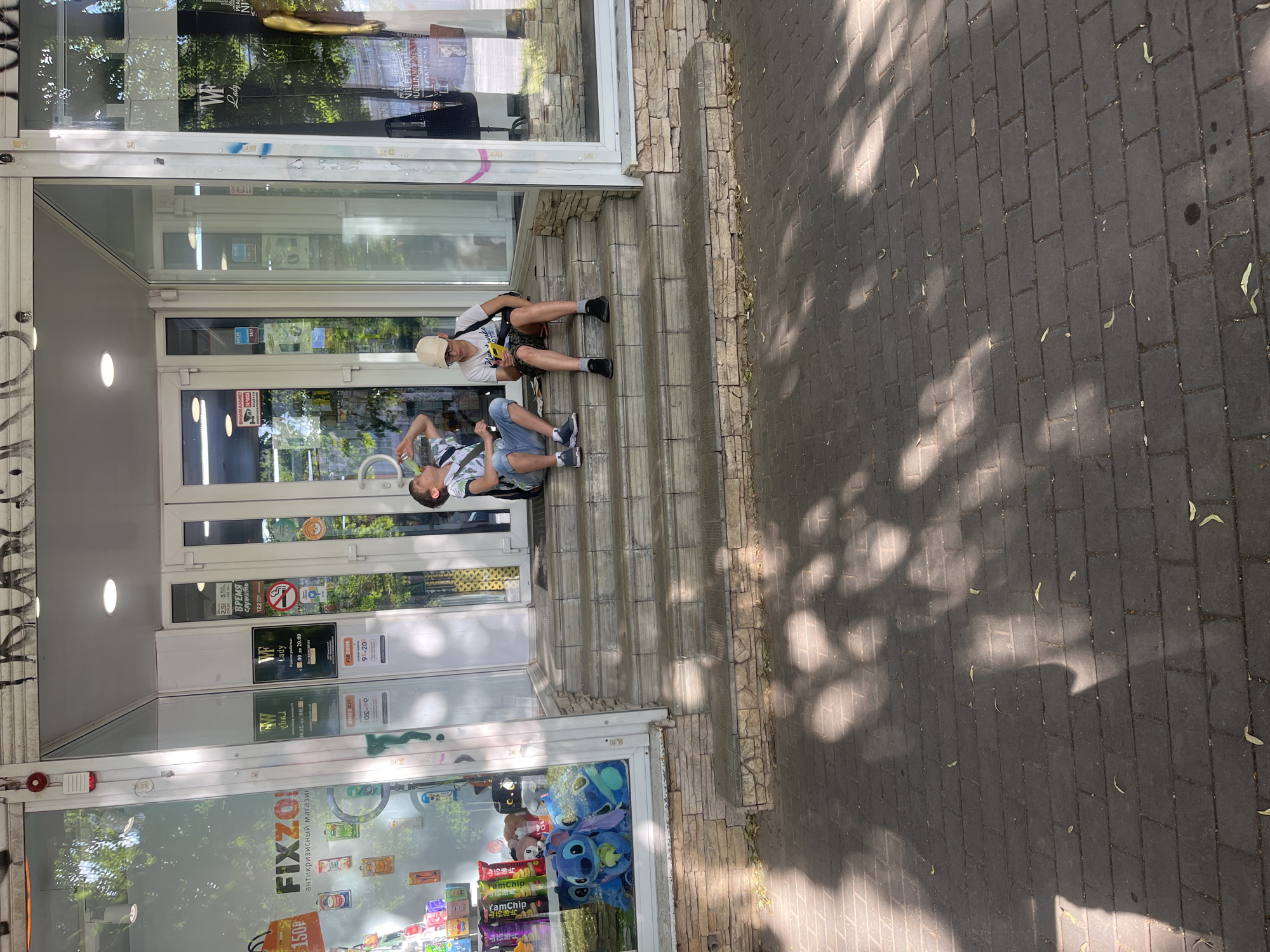 Two young individuals are seated on outdoor steps in front of a store. The person on the upper step, wearing a white cap and a white t-shirt with a black and white patterned trim, holds a yellow object. They are wearing shorts and black shoes. The person on the lower step, wearing a white t-shirt with a similar patterned trim and a green and black patterned t-shirt over it, is wearing blue jeans and black shoes. Both individuals appear relaxed.
The store's entrance features large glass doors and windows, with signage indicating it is a "FIXZO!" "антикризисный магазин" (anti-crisis store). Other visible text includes "WF Lady" with operating hours "с 8.00 до 20.00", "FIXZONE 9-20", and a "24 ЧАСА 24/7" sign. The store appears to sell a variety of items, including plush toys visible through the window, such as a Stitch character.
The scene takes place during the daytime, with dappled sunlight casting shadows from trees onto the brick pavement in front of the steps. The overall atmosphere is casual and relaxed, suggesting a warm, sunny day. The location is identified as Voronezh, Russia.