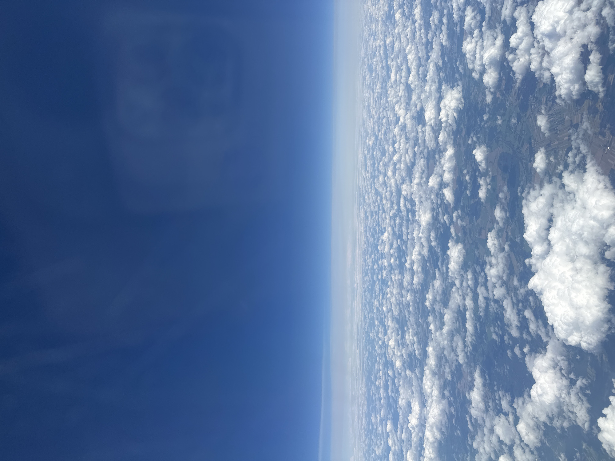 An aerial view shows a vast expanse of fluffy white clouds covering the landscape. The clouds are scattered across the scene, with some appearing larger and more defined than others. Peeking through the gaps in the clouds, faint outlines of fields and possibly some scattered buildings can be observed, suggesting a rural or semi-rural area. The sky above is a clear, deep blue, creating a stark contrast with the white clouds below. The image appears to be taken from an aircraft, as indicated by the high vantage point and the horizon line. The location of Клейнікаўскі сельскі Савет, Belarus, is a rural administrative division, which aligns with the observed landscape.