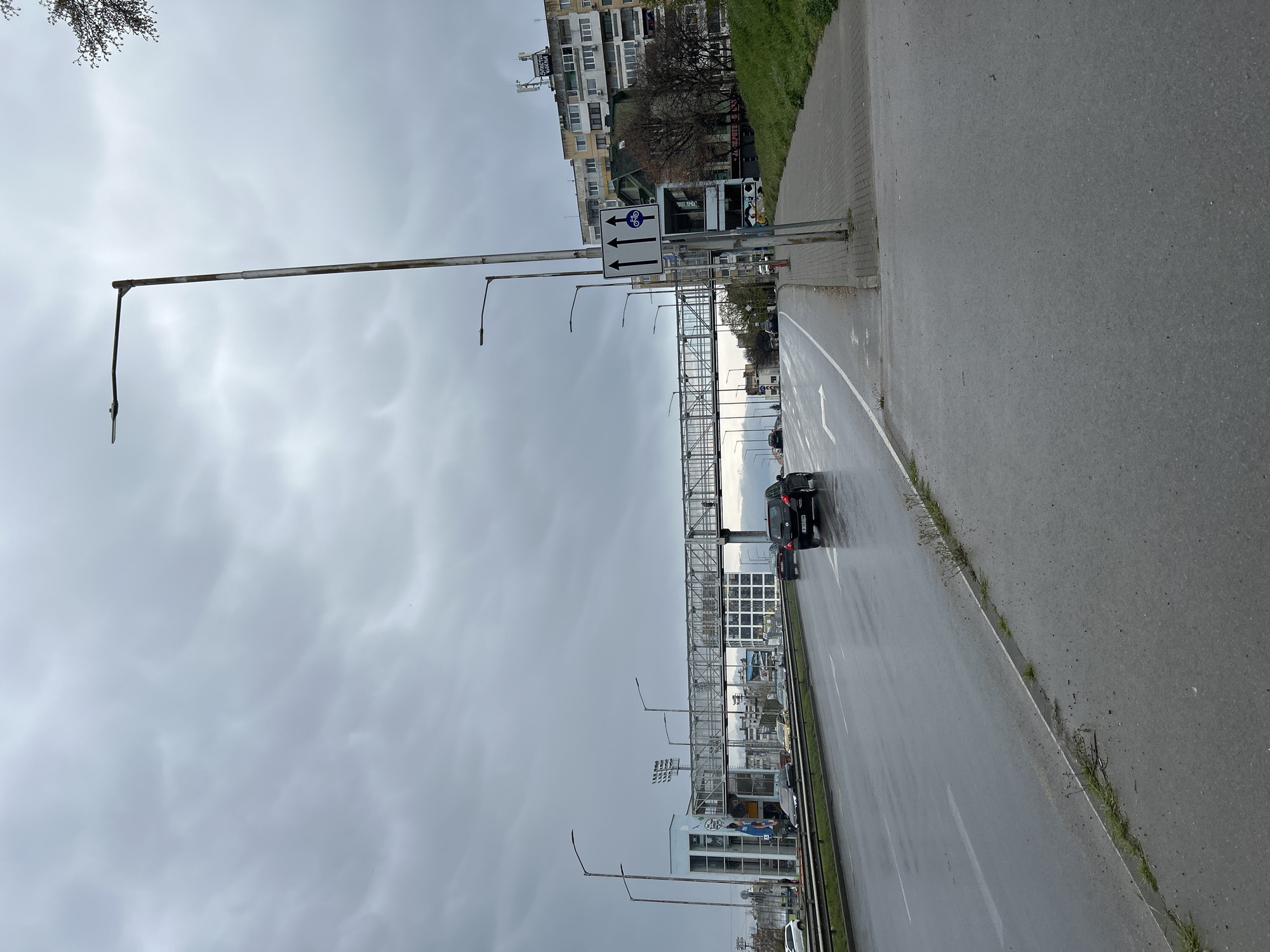 The image depicts a street scene in Varna, Bulgaria, under a cloudy, overcast sky. The street is wet, likely due to recent rain. There are several cars on the road. A pedestrian overpass spans the road. A road sign is visible that has a bicycle lane, directing to go straight or turn left. Buildings are visible in the background, including some with satellite dishes. Grass grows along the sidewalk and median. Streetlights line the roadway. The overall scene evokes a sense of urban life on a rainy day.