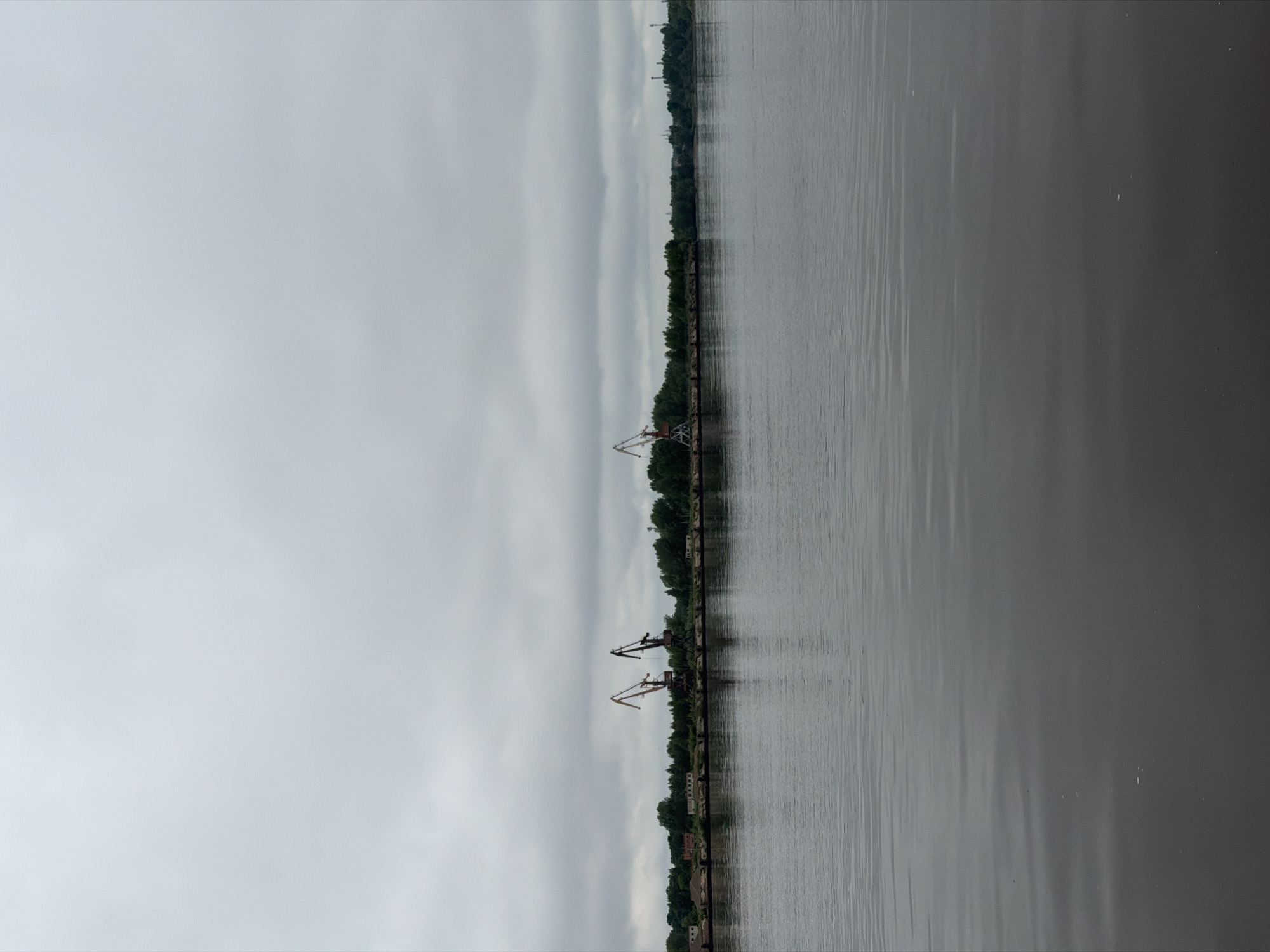 The image, when viewed horizontally, displays a wide river or large body of water under an overcast sky. The scene is divided horizontally into three main sections: a vast, cloudy sky, a distant shoreline with industrial structures and vegetation, and a broad expanse of water in the foreground.
The sky is a uniform light grey, indicating an overcast or heavily clouded day. Subtle bands of lighter and darker grey clouds are visible, but no direct sunlight penetrates. The water in the foreground is dark and calm, with minimal ripples, reflecting the muted light from the sky. Its color is a deep grey or brownish-grey.
Along the distant bank, a narrow strip of land is visible, densely covered with green trees and foliage. Several large, dark industrial cranes rise above the tree line. There are at least four visible cranes, positioned along what appears to be a pier or industrial waterfront. These cranes are silhouetted against the sky and water, suggesting a port or heavy industrial area. Some faint, darker structures are also barely discernible behind the trees and near the water's edge on the far bank. A long, dark line, possibly a dock or retaining wall, runs parallel to the shore along the water.
No specific activity or individuals are visible. The scene conveys a quiet, perhaps somber, atmosphere due to the overcast conditions. There is no visible text within the image. Given the context, this industrial river scene is located in Chaikovsky, Russia.