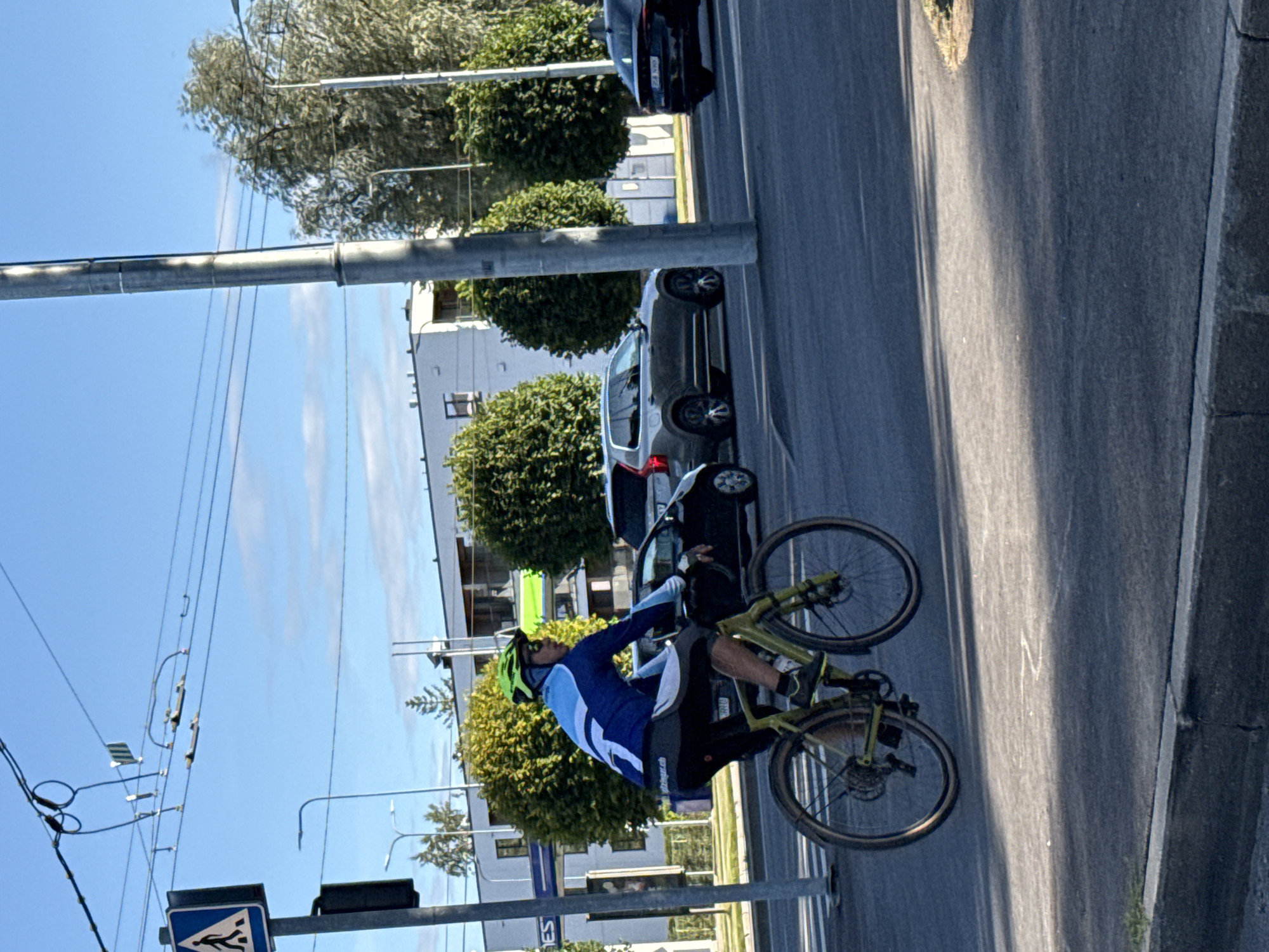 The image captures a street scene in Tallinn, Estonia, with a cyclist in the foreground. The cyclist, wearing a blue and white long-sleeved jersey, black shorts, and a green helmet, is riding a yellow-green bicycle. The bicycle has black tires with silver spokes and appears to be a gravel or cyclocross bike. The cyclist's left leg is extended, and the front wheel is slightly elevated, suggesting the cyclist might be performing a small jump or maneuver.
In the background, traffic is visible on the road. A dark-colored SUV, possibly a Volvo XC90, is stopped or moving slowly in the right lane. Further down the road, a black sedan is also present. Buildings with white facades and dark windows are visible behind the trees. The street is paved, and there are power lines overhead, supported by utility poles. One of the poles, a prominent grey cylindrical structure, stands to the right of the cyclist. A pedestrian crossing sign with a blue background and a white pedestrian symbol is visible on the left side of the image, mounted on a black pole. The sky is blue with scattered white clouds, indicating daytime. The lighting suggests it's a sunny day with some shadows cast on the road. The environment appears to be an urban or suburban intersection.