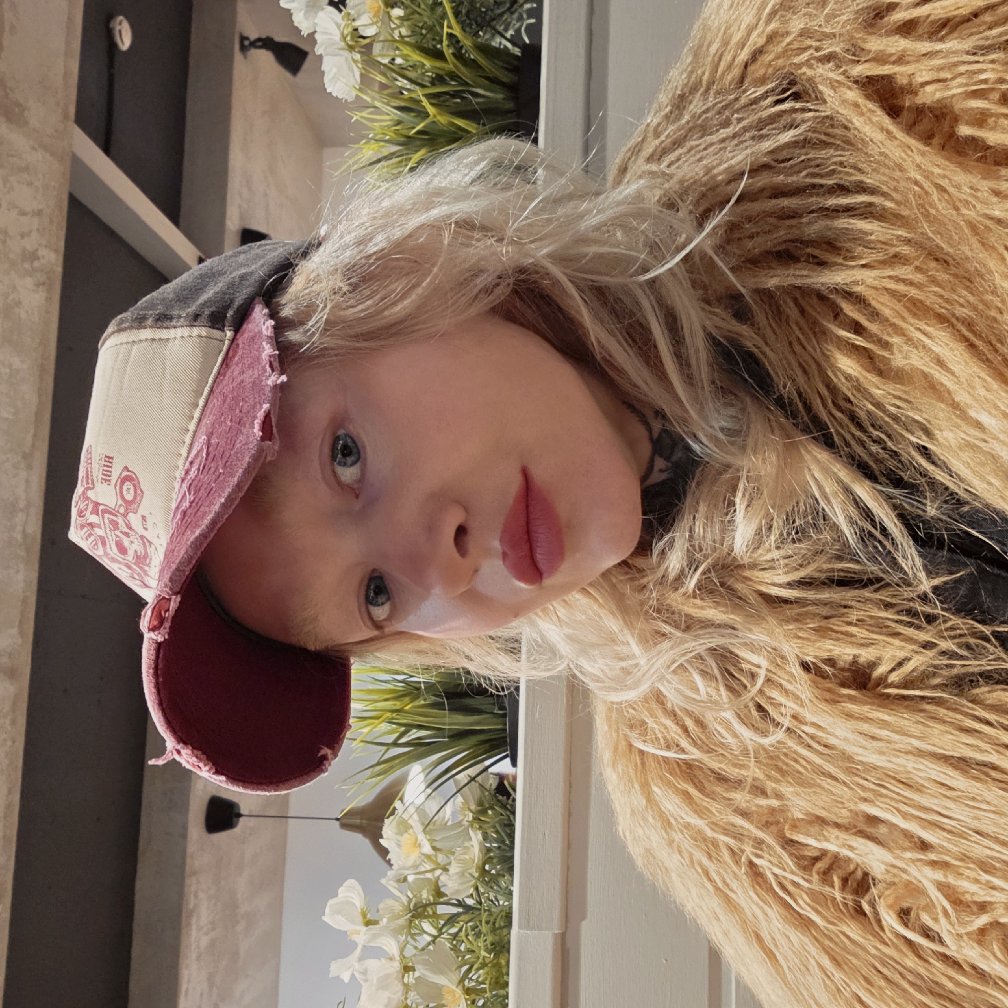 A fair-skinned person with blonde hair wearing a distressed baseball cap and a shaggy tan jacket poses for a selfie. The cap is a mix of colors with a burgundy brim, cream-colored front panel displaying a red tractor graphic, and a dark gray upper section. The person has light blue eyes and pink lips. The background features a plant arrangement of green leaves and white flowers in a white rectangular container. Above the planter, the corner of a shelf or wall is visible, with a dark gray element above it. The location is impossible to determine from the provided information.