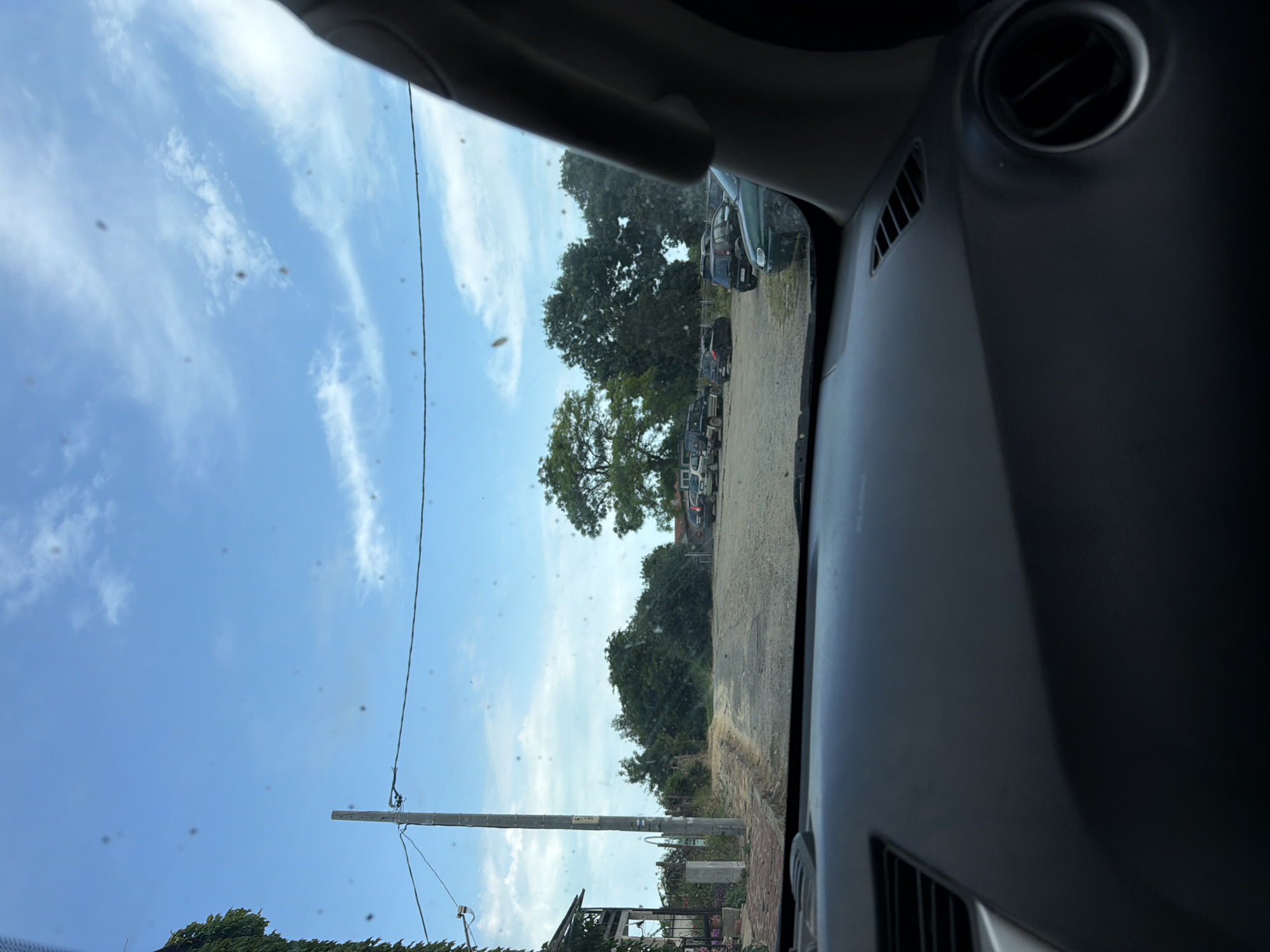 The image presents an exterior scene viewed through a car's windshield, which exhibits numerous small, dark spots, likely dust or debris. The lower portion of the frame is occupied by the dark gray dashboard of the vehicle, featuring a rectangular air vent on the left and a circular air vent with silver trim on the far right.
Outside the vehicle, an unpaved, dirt path extends into the distance. To the left, a building partially covered in green climbing vegetation is visible, alongside a light gray utility pole with horizontally strung electrical wires. Further ahead, dense green trees form a natural backdrop. To the right of the path, a dirt parking area contains multiple parked vehicles, including a dark SUV, a dark hatchback, a black SUV/crossover, and a dark green sedan positioned in the immediate foreground on the right. A line of mature trees delineates the far side of the parking area. The sky above is clear blue with scattered white clouds. The scene depicts a static, daytime environment consistent with a rural or semi-rural area in Rezovo, Bulgaria. No people, active movements, or interactions are visible.