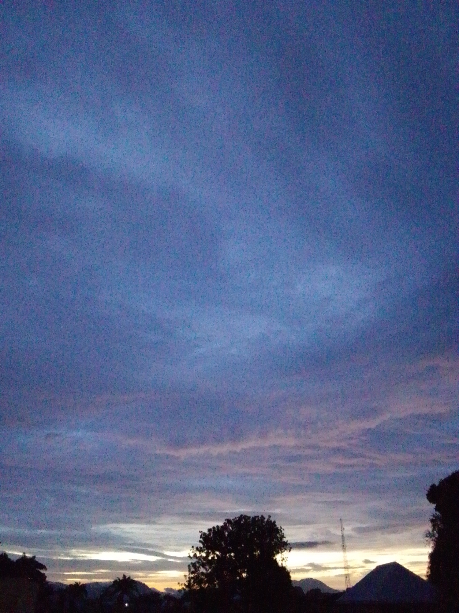 This is a dusk or dawn photograph taken in Serti, Nigeria. The sky dominates the upper two-thirds of the image, displaying a gradient of deep blue and purple hues with streaks of lighter clouds tinged with pink and orange near the horizon. The lower portion of the image shows a silhouetted landscape. In the foreground, on the right, the dark silhouette of a tree is visible. To the left of center, a large tree with a full canopy is also silhouetted. In the distance, the silhouettes of hills or mountains are apparent, with a prominent, conical structure (possibly a building or a natural formation) on the right side. A tall, thin tower, likely a communication tower, stands to the left of the conical structure. The horizon line is low, with the sky being the primary focus. The overall impression is one of a natural landscape at twilight.