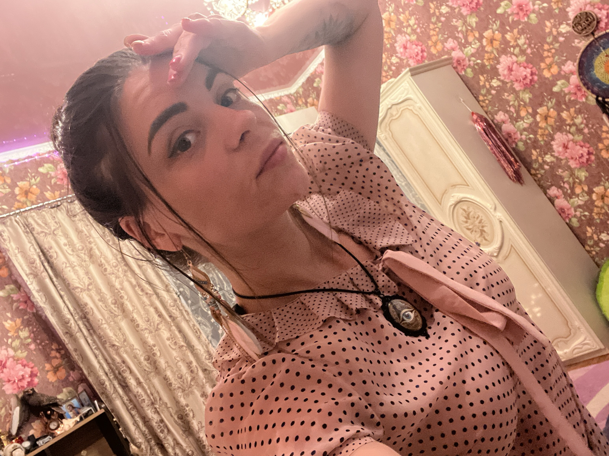 In this indoor, close-up, selfie-style photograph, a young woman with dark hair is the central subject. She is wearing a pink, polka-dotted blouse with a ruffled collar and a necklace featuring a dark pendant with an eye design. Her right hand is raised to her forehead, with fingernails painted red. Her expression appears thoughtful or perhaps slightly pensive.
The background reveals a bedroom setting. To the right, a portion of a white ornate wardrobe or cabinet is visible, against a floral-patterned wallpaper featuring pink and orange flowers. To the left, a light-colored, patterned curtain is drawn back, partially obscuring a shelf with various decorative items, including framed pictures and a clock. A string of pink LED lights is also visible in the upper left corner, suggesting the time of day might be evening or night, or that the lights are used for ambiance.
The overall mood of the image is intimate and personal, with the focus on the woman's face and upper body. The lighting appears to be a mix of ambient room light and possibly artificial light from the LED strip. No text is visible in the image. The location context of Krasnoe-na-Volge, Russia, is provided as metadata.