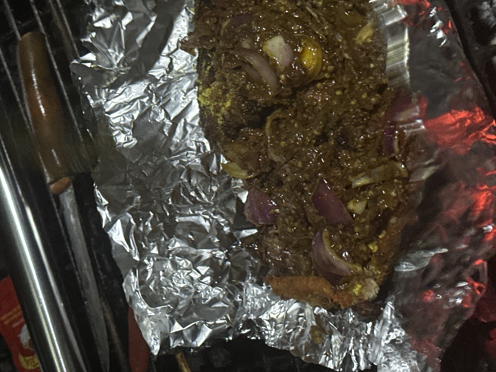 A close-up, overhead shot shows food being cooked on a grill. The food, which appears to be meat with onions and spices, is wrapped in aluminum foil. The foil is crumpled and shiny, reflecting the heat from the coals below. Red embers glow beneath the foil, indicating an active fire. A metal rod, likely part of the grill, is visible at the top of the image, along with a portion of what looks like a red and yellow package. The scene suggests an outdoor cooking environment, possibly in Jos, Nigeria, where grilling is a common practice. There are no people visible in the frame.