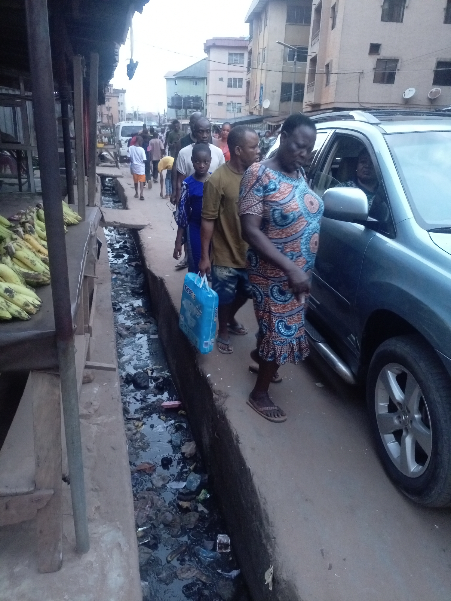 This street-level photograph captures a bustling urban scene in Onitsha, Nigeria, showing a mix of pedestrian activity, local commerce, and a roadside environment.
In the foreground, a group of people is seen walking along a narrow pathway. A woman in a vibrant, patterned blue, orange, and white dress and sandals walks alongside a dark-colored SUV. Behind her, a man in a brown shirt and patterned shorts carries a bright blue shopping bag with partial text "KISSK S" visible. Further back, other pedestrians, including a young boy in a blue top and several adults, navigate the path. The driver of the SUV is partially visible through the vehicle's window.
On the left side of the image, wooden market stalls display numerous bunches of green plantains or bananas. Adjacent to these stalls and running along the pedestrian path is an open, concrete gutter filled with stagnant water and a considerable amount of trash, including plastic bags and other debris.
The background reveals multi-story residential or commercial buildings lining the street, some equipped with satellite dishes, indicating a densely populated urban area. A white van is also visible further down the street. The sky appears overcast, casting a soft, diffused light over the scene, suggesting either early morning, late afternoon, or a cloudy day. The overall image portrays daily life in a busy city, characterized by people moving about, local trade, and the visible presence of urban infrastructure and waste.
