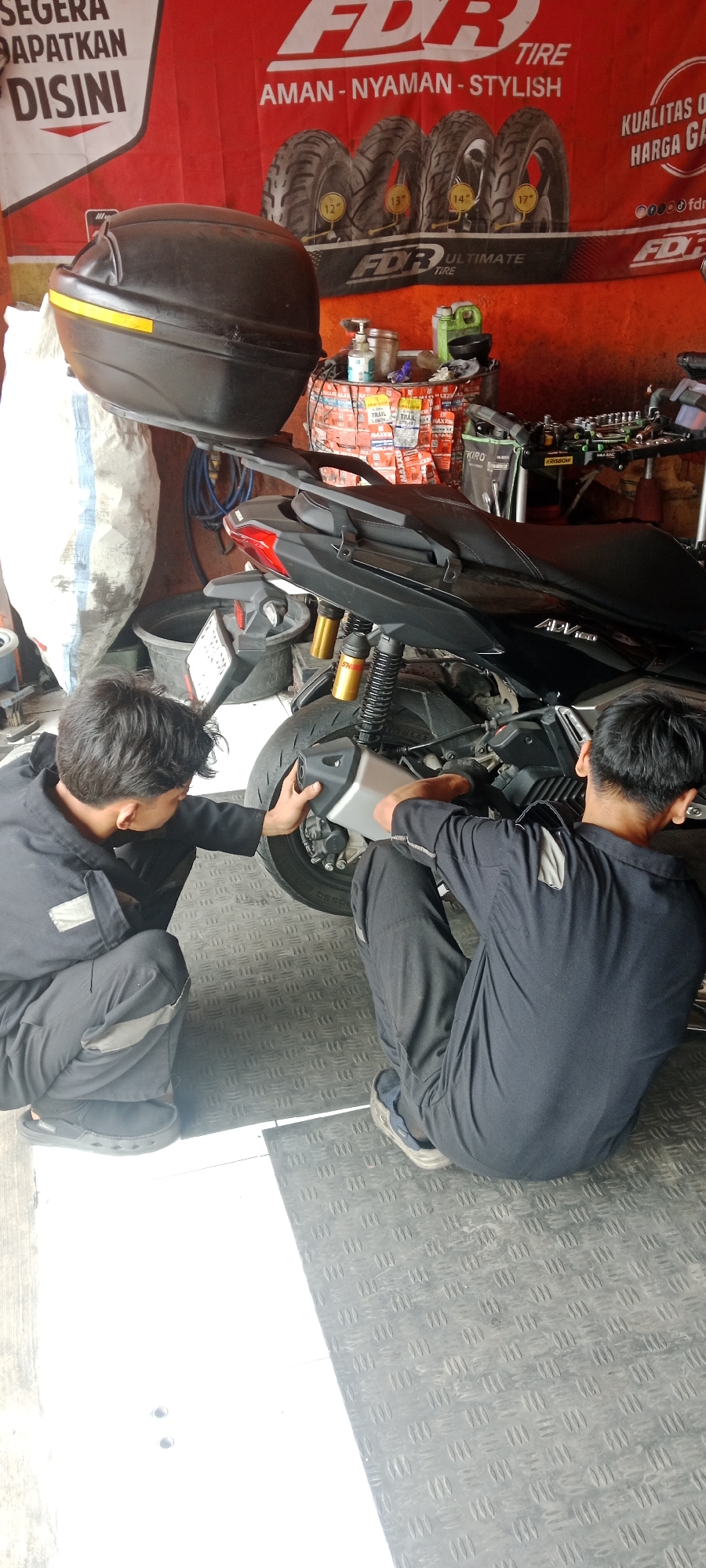 In a workshop in Tanjakan, Indonesia, two individuals in dark grey work uniforms are crouched down, attending to the exhaust pipe of a black scooter. The scooter is positioned on a textured metal floor. One individual, on the left, is wearing black sandals, while the other, on the right, wears grey sneakers. In the background, a red banner with "FDR TIRE" prominently displayed is visible, along with several tires and other workshop equipment. A black motorcycle luggage box is also present in the scene.