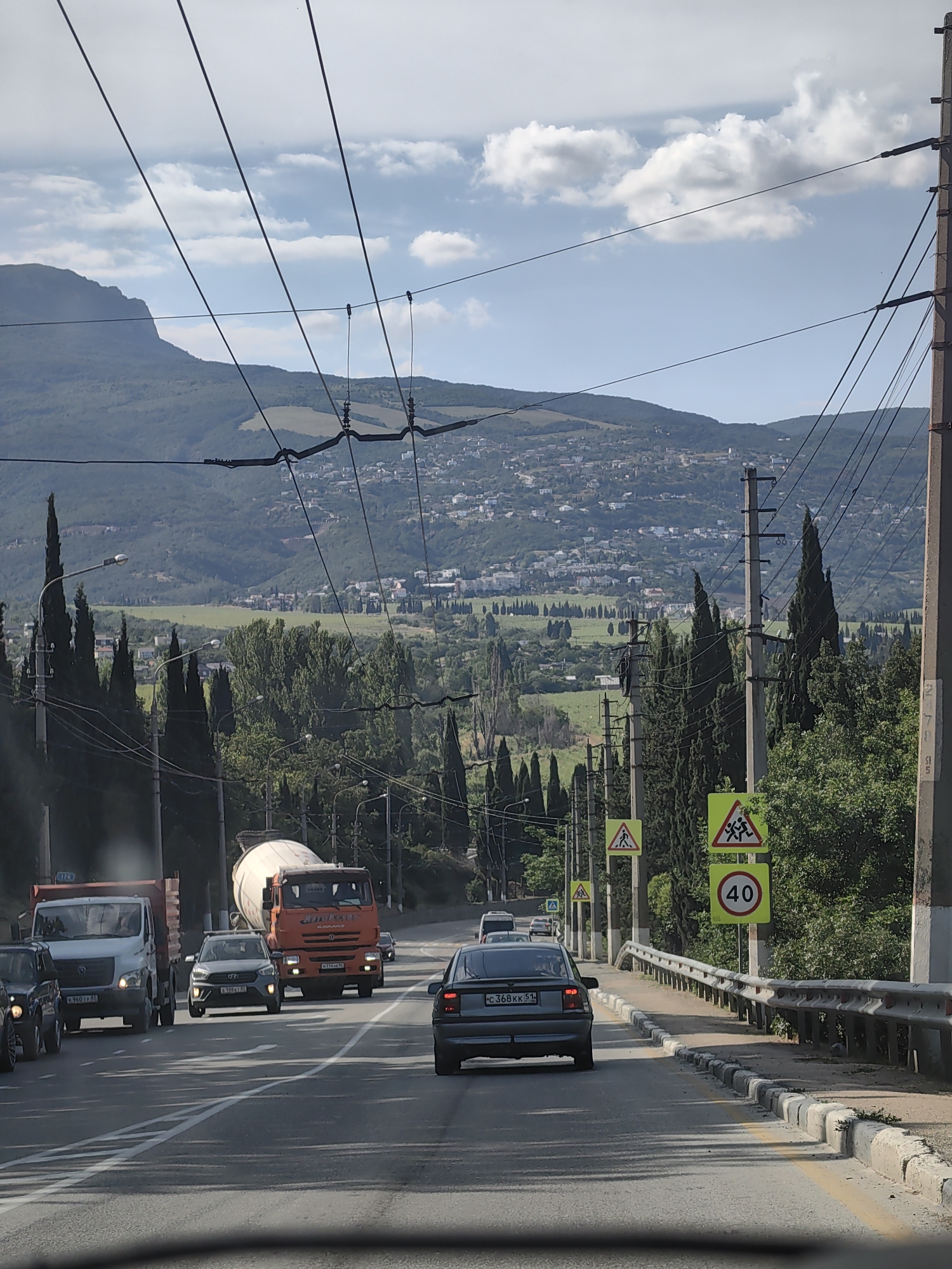 The image shows a road in Alushta, Ukraine, with cars and a cement truck driving on it. In the background, there are hills with houses and trees. The sky is blue with some clouds. There are power lines running across the scene. There are also several road signs visible, including a speed limit sign of 40. The road is asphalt and appears to be in good condition. The cars are of various makes and models. The cement truck is orange and white. The environment appears to be a sunny day with moderate traffic.