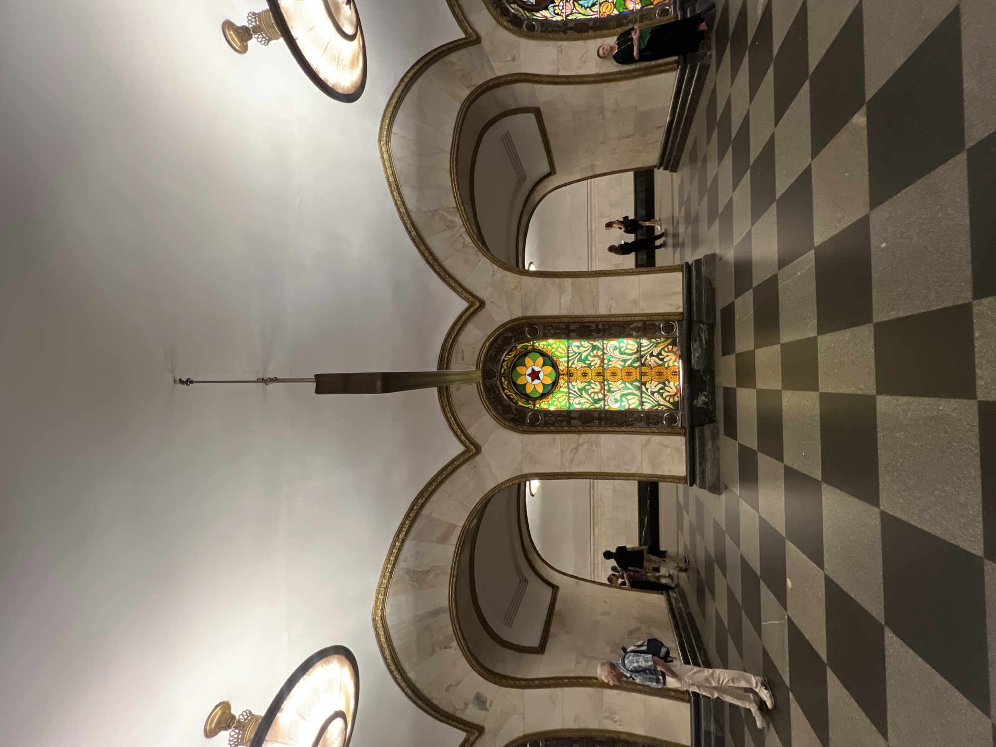 The image shows an interior view of a building with a distinctive architectural style. The ceiling is white, and there are ornate light fixtures in the top and bottom corners of the image. Arched openings with gold-colored trim and marble-like details are visible, leading to other areas within the building. A stained-glass window is positioned centrally in the view, featuring a colorful design. People are present in the image, appearing in the arched openings and on the floor. The floor has a checkered pattern in dark and light shades. The location is unidentified; therefore, city information is unavailable.
