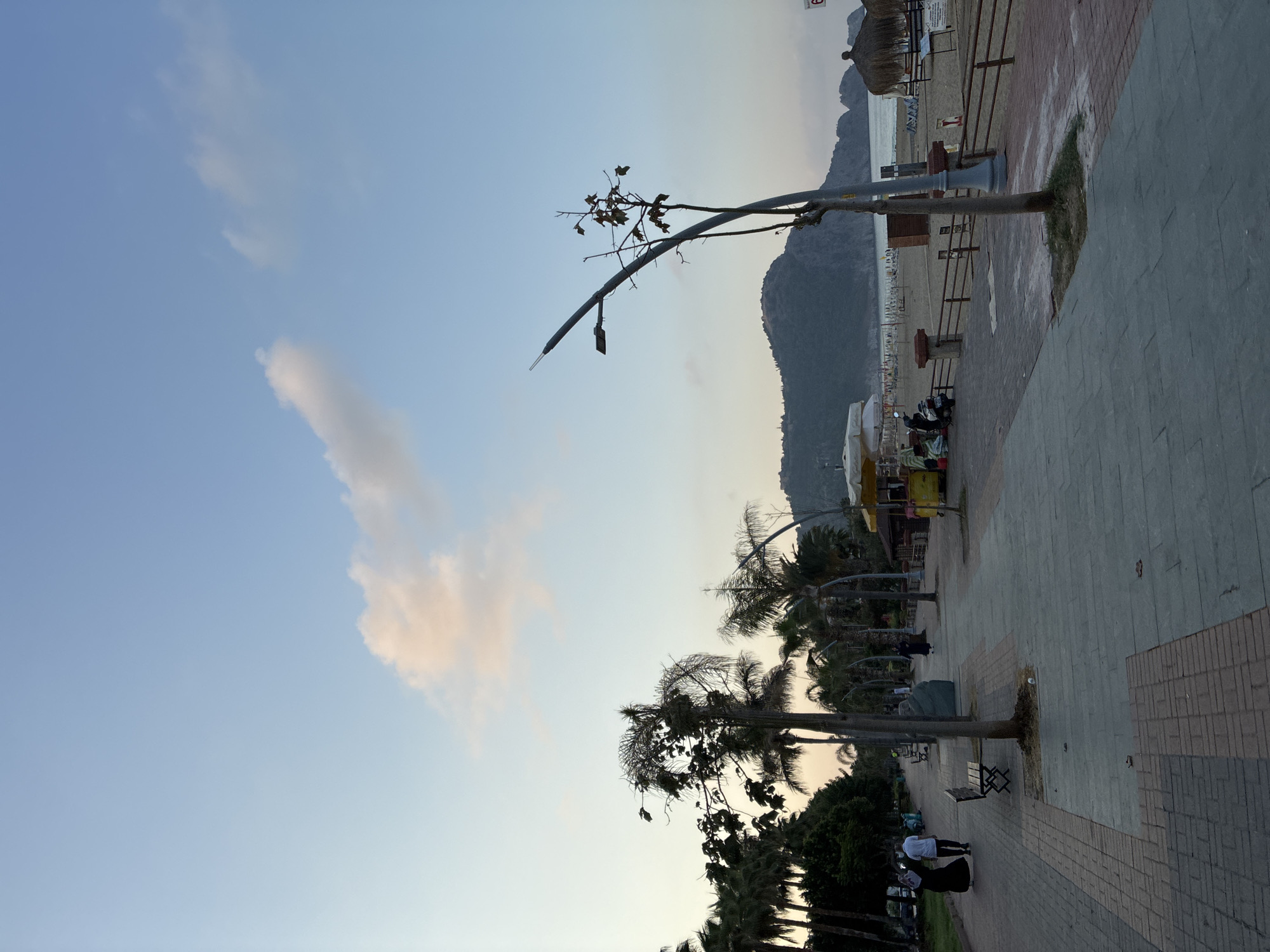 The image depicts a seaside promenade in Alanya, Turkey, at dusk. A paved walkway, composed of grey stone slabs and red brick borders, runs along the left side of the frame, bordered by palm trees. Several people are walking on the promenade, including a couple dressed in traditional Islamic clothing, with the woman in a black abaya and the man in a white t-shirt and dark pants. Benches are placed along the walkway. Further down, a yellow food cart is visible, with a motorcycle parked nearby.
To the right of the promenade is a sandy beach with rows of beach chairs and umbrellas. In the background, a low, tree-covered mountain range rises from the sea, its silhouette softened by the twilight. The sky is a clear blue with a few wispy clouds tinged with pink and orange from the setting sun. A tall lamppost with a modern design stands prominently in the middle ground, its arm angled over the promenade. The overall atmosphere is tranquil, characteristic of an evening stroll along the coast.