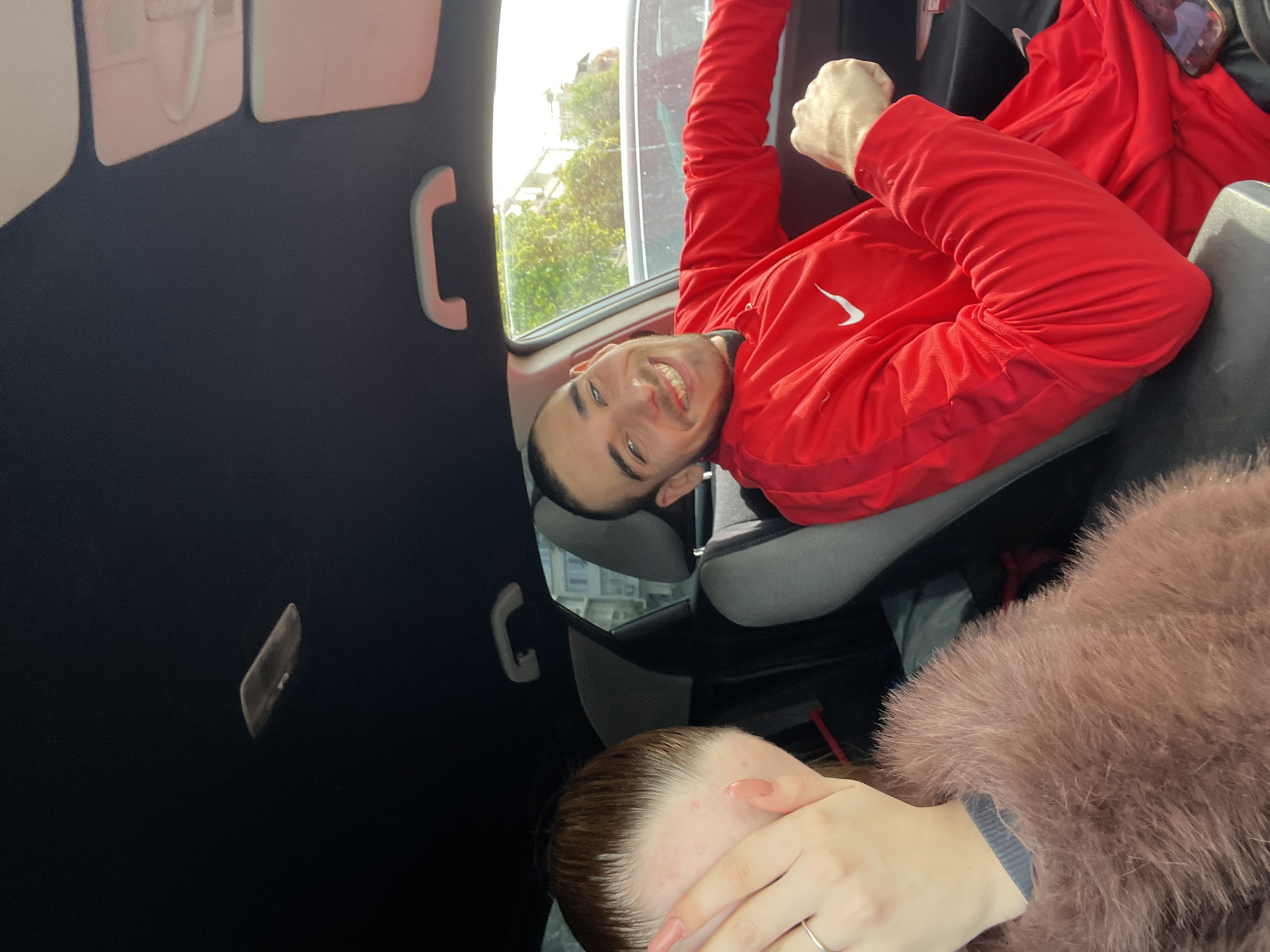 A medium shot captures a man in a red athletic jacket posing inside a vehicle. He is smiling and has his fist clenched. To the side and below him is a person wearing a fur coat and covering their face. Through the window, a building and trees are visible. The location is Varna, Bulgaria.