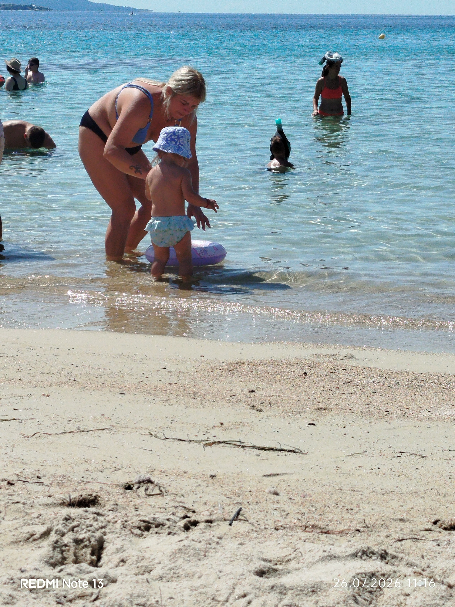In Sithonia Municipal Unit, Greece, on a sunny day, several individuals are enjoying the shallow, clear turquoise water near a sandy beach. In the foreground, a woman wearing a blue bikini top and black bottoms is crouched down, interacting with a toddler. The toddler, wearing a blue and white patterned diaper and a matching hat, stands in the water with a pink inflatable ring behind them. Further back, a person in a bright orange bikini is standing in the water, wearing a snorkel mask on their head. Another person, mostly submerged, also appears to be wearing snorkeling gear. In the distance, the coastline with hills is visible under a bright blue sky. The beach itself is composed of light-colored sand with scattered shells and debris. The timestamp "26.07.2026 11:16" and "REDMI Note 13" are superimposed on the bottom of the image.