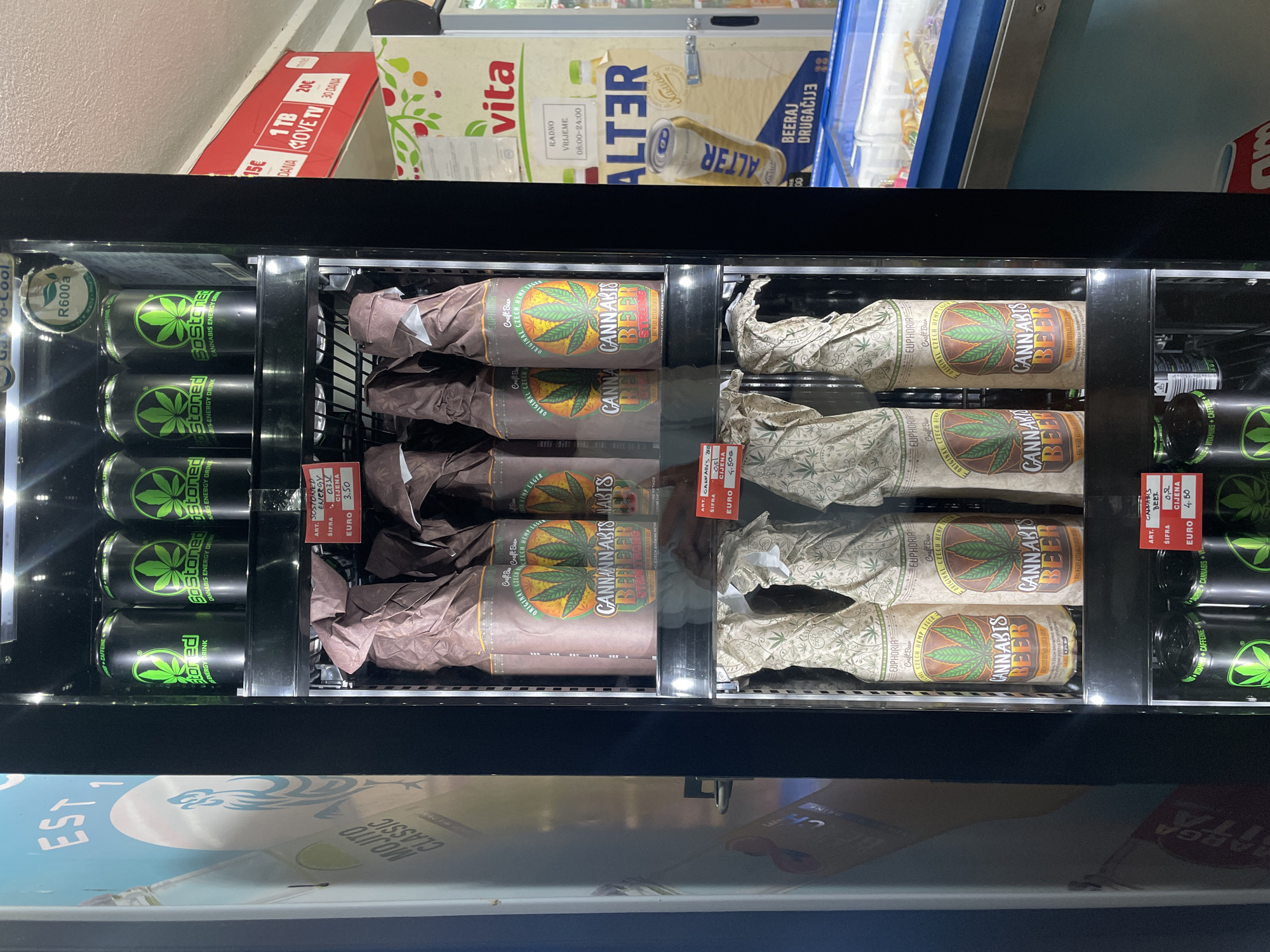 An interior view depicts a black, glass-fronted commercial refrigerator unit displaying various cannabis-themed beverages, positioned in what appears to be a convenience store or market in Unknown, Unknown.
The top shelf of the refrigerator contains four black cans labeled "GO STONED CANNABIS ENERGY DRINK" featuring a prominent green cannabis leaf logo. A red price tag indicates "0.33L" and a "CIJENA" (price) of "EURO 3.50".
The middle shelf holds multiple glass bottles, largely wrapped in crumpled brown paper. Visible labels show a green cannabis leaf, "CANNABIS BEER STRONG," and "ORIGINAL CZECH HEMP LAGER / CRAFT BEER." Another red price tag, partially obscured, shows "0.5L" and "EURO 4.50" for "CANNABIS BEER."
The bottom shelf also displays glass bottles, similarly wrapped, but in a lighter-colored paper patterned with small cannabis leaves. Visible labels again state "EUPHORIA CANNABIS BEER" and "ORIGINAL CZECH HEMP LAGER."
To the left of the cooler, a light blue wall or large advertisement displays white text including "EST 1" and "MOJITO CLASSIC."
To the upper right of the cooler, a red rectangular sign advertises "1 TB MOVE TV" for "20€ / 30 DANA" (30 days). Below this, a cream-colored poster features "vita" and an "ALTER" beer can, stating "RADNO VRIJEME 08:00-24:00" (Working Hours 08:00-24:00) and "BEERAJ DRUGAČIJE" (Beer Different).
In the lower right foreground, a blue refrigerated display case containing various packaged items is partially visible.
The scene is well-lit by internal refrigerator lighting and ambient store lighting. No individuals are present in the image. Text is visible in both English and what appears to be Croatian or Serbian.