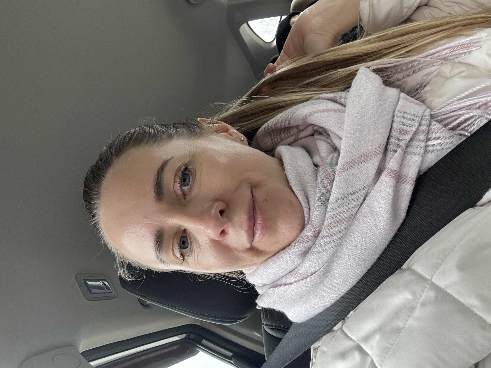 A woman with fair skin, blue eyes, and blonde hair is sitting inside a car. She is wearing a light pink and white scarf and a light-colored quilted jacket. A black seatbelt crosses her torso. The interior of the car is visible, including the headrest of the seat, the car's ceiling, and a glimpse of the outside through the window. The photograph has been taken at an angle.
There is no information present in this image to indicate the location is Chaikovsky, Russia.