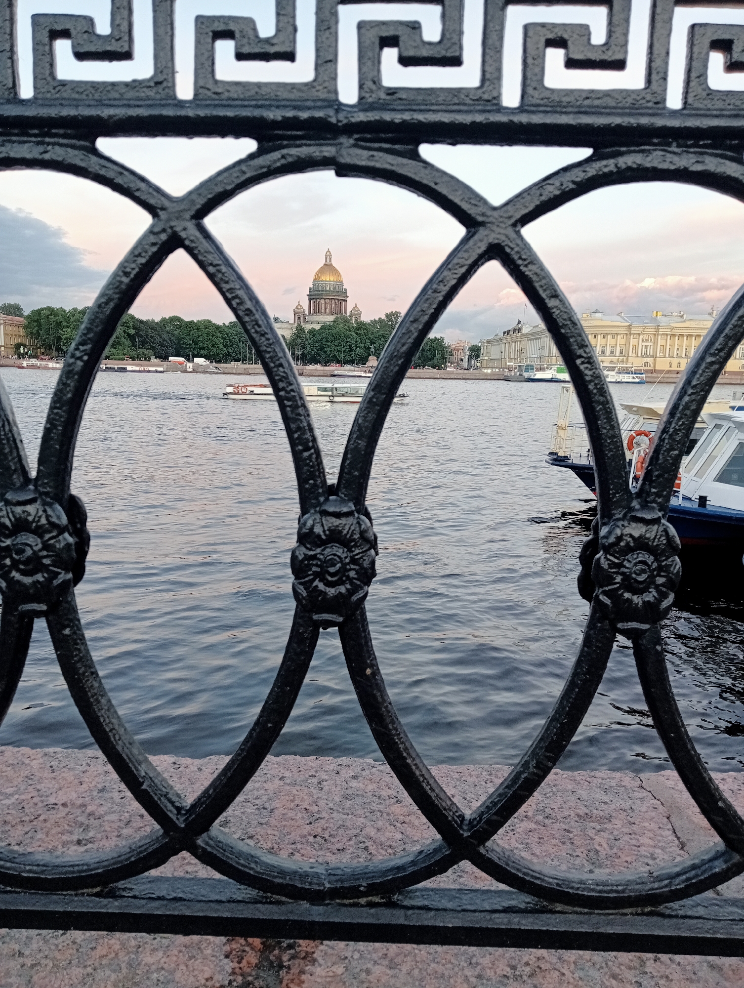 A river with several boats is visible in the middle ground. In the background, a large building with a golden dome stands prominently, flanked by other buildings and trees. The foreground features a decorative black metal fence. The sky above is overcast with pinkish hues, suggesting either dawn or dusk. This view is likely from the Neva River embankment in Saint Petersburg, Russia, with St. Isaac's Cathedral being the prominent golden-domed structure.