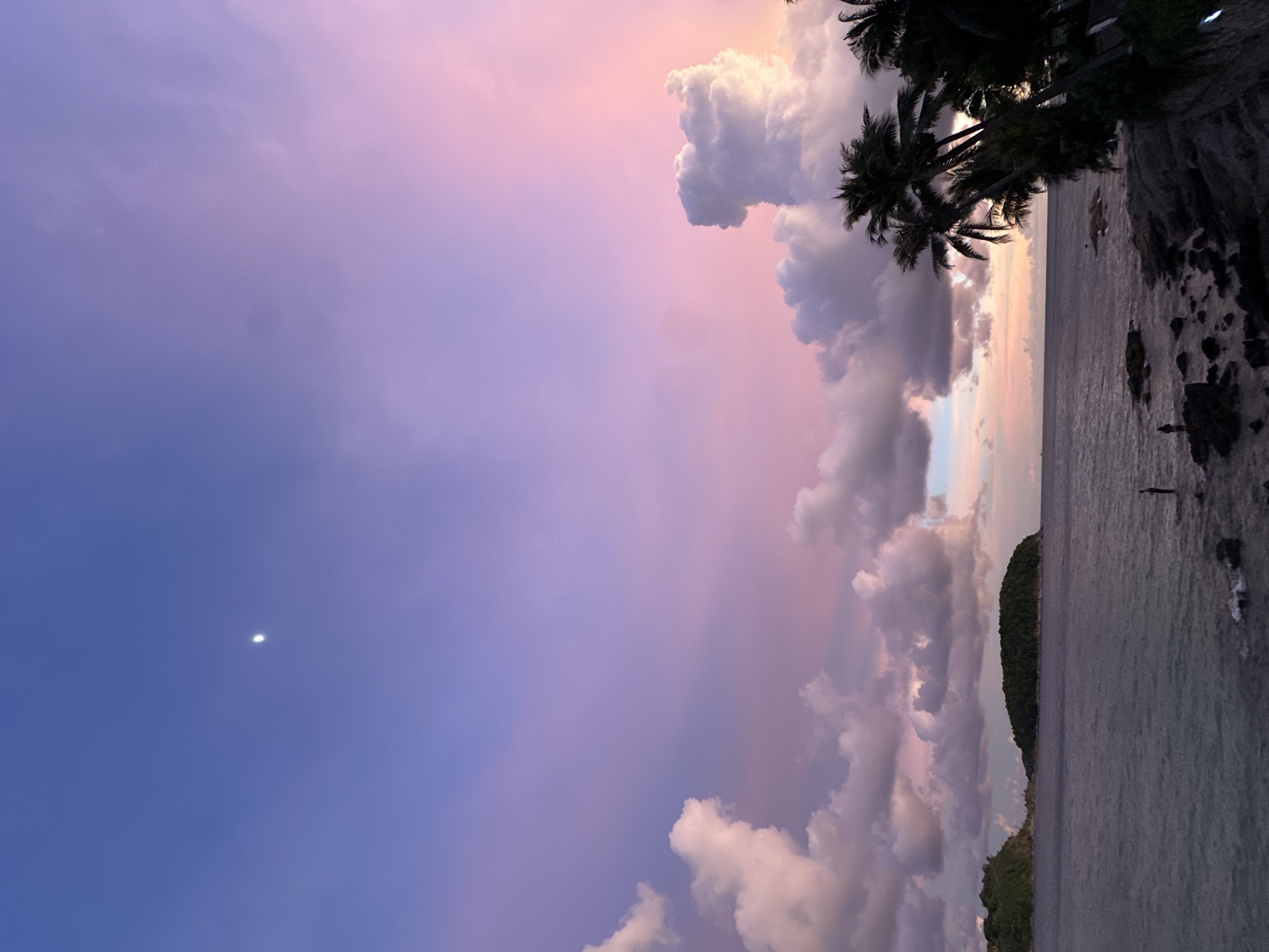 The image depicts a serene coastal scene at twilight in Rawai, Thailand. The sky is a canvas of soft purple and pink hues, transitioning into a deep blue at the lower left, where a crescent moon is visible. Fluffy clouds, tinged with pink and white, dominate the right side of the sky, catching the last rays of the setting sun. Silhouetted palm trees, dark against the colorful sky, frame the upper right corner of the scene.
The foreground is a rocky shoreline meeting the calm ocean. The water is a muted gray, reflecting the dim light. Scattered rocks emerge from the water near the shore. On the right edge of the image, a lone figure stands in the shallow water, their dark silhouette a small contrast against the vast expanse of sea and sky. In the distance, a lush green island rises from the sea, its shape a gentle curve against the horizon.
The overall mood is peaceful and tranquil, characteristic of a sunset by the sea. The soft colors and stillness of the scene evoke a sense of calm and reflection. The image captures a moment of natural beauty as day transitions into night. There is no visible text in the image.