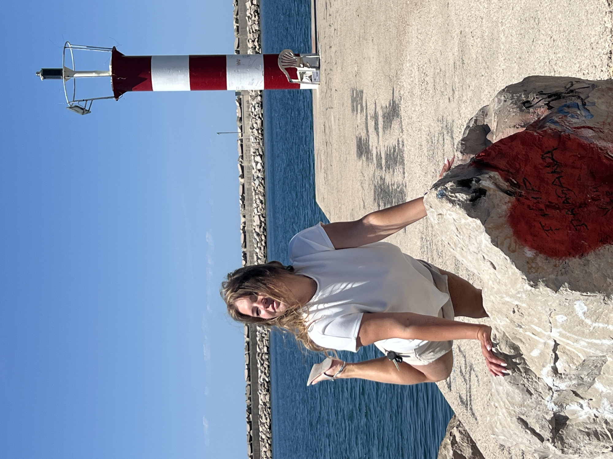 The image depicts a bright, sunny day at a coastal location, identified as Bar, Montenegro. The central subject is a young woman with long, wavy blonde hair, wearing a white t-shirt and light-colored shorts. She is shown in an active pose, seemingly climbing or posing on a large, irregularly shaped rock in the foreground. Her left hand grips the rock, and she looks towards the viewer with a pleasant, perhaps slightly playful, expression. Her fingernails are painted white.
The rock on which she poses has visible patches of red, black, and blue graffiti, though any specific text or symbols are illegible. In the upper left portion of the frame, a distinctive red and white striped navigational beacon stands. Faint, illegible black text is visible on one of the white stripes of the beacon. Next to the beacon, a small, white, shell-shaped decorative chair is present.
The background features calm, deep blue water, characteristic of a clear day at sea. A concrete pier or breakwater extends across the midground, providing a barrier between the foreground and the water. Further in the distance, a rocky coastline or another breakwater is visible. The sky is a vibrant, clear blue, suggesting midday or early afternoon conditions. The overall scene conveys a sense of outdoor activity and leisure in a scenic maritime setting.