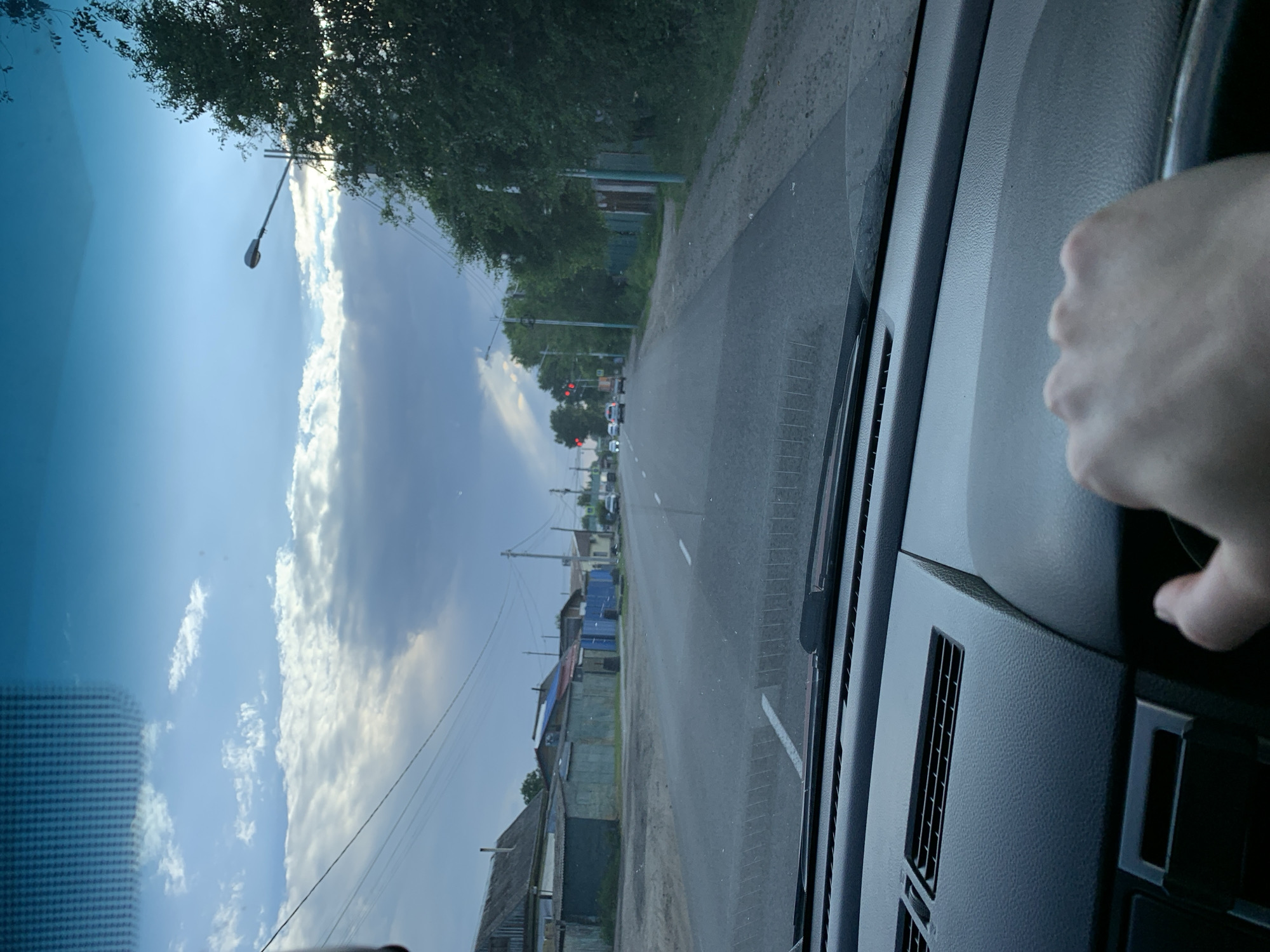 The image depicts a view from the driver's seat of a car looking forward through the windshield. A person's right hand is visible on the steering wheel. The car is on a paved road with a white lane marking. To the left of the road, there are houses with corrugated metal roofs and fences, and utility poles with wires. To the right, there are trees and more houses. In the distance, traffic lights are visible, showing red. The sky is cloudy with sunlight breaking through in patches. The scene appears to be in a residential area of a city.