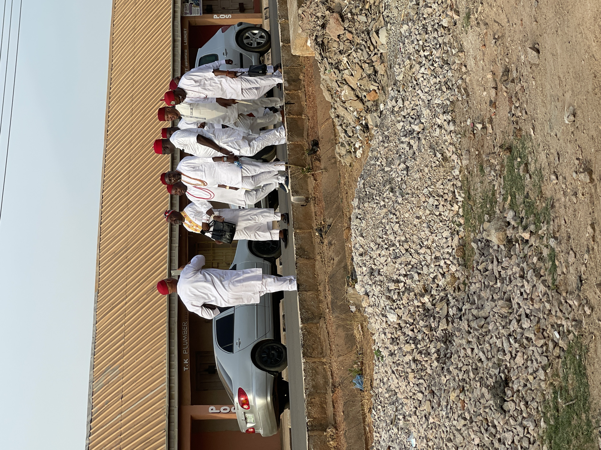The image captures a group of approximately ten men, predominantly dressed in white traditional Nigerian attire, standing in a line along the side of a building. Most of the men wear distinctive red caps, often associated with cultural traditions in parts of Nigeria, such as Igbo culture. Several individuals also display elaborate bead necklaces or chains, some featuring red and white beads.
The scene unfolds during daylight hours under a bright sky, suggesting good weather conditions. The men are positioned on a paved surface next to a light-colored building with a distinctive corrugated metal roof. Adjacent to the paved area, the ground transitions into an unpaved, uneven terrain composed of gravel, small stones, and patches of dirt with minimal green vegetation, typical of a roadside or undeveloped area.
Two cars are visible near the building: a silver sedan and a lighter-colored hatchback, both parked or stopped close to the structure. The building itself appears to house various commercial establishments, as indicated by visible text on its facade. Clearly discernible signs include "T.K PLUMBER" written vertically, accompanied by a phone number "0816462428". Other partially visible words include "POS" (likely Point of Sale) and "LAUNDRY".
The men appear to be gathered or waiting, possibly for a formal event, a social occasion, or engaging in conversation. Their composed postures and traditional attire suggest a cultural or communal gathering in Narayi, Nigeria.